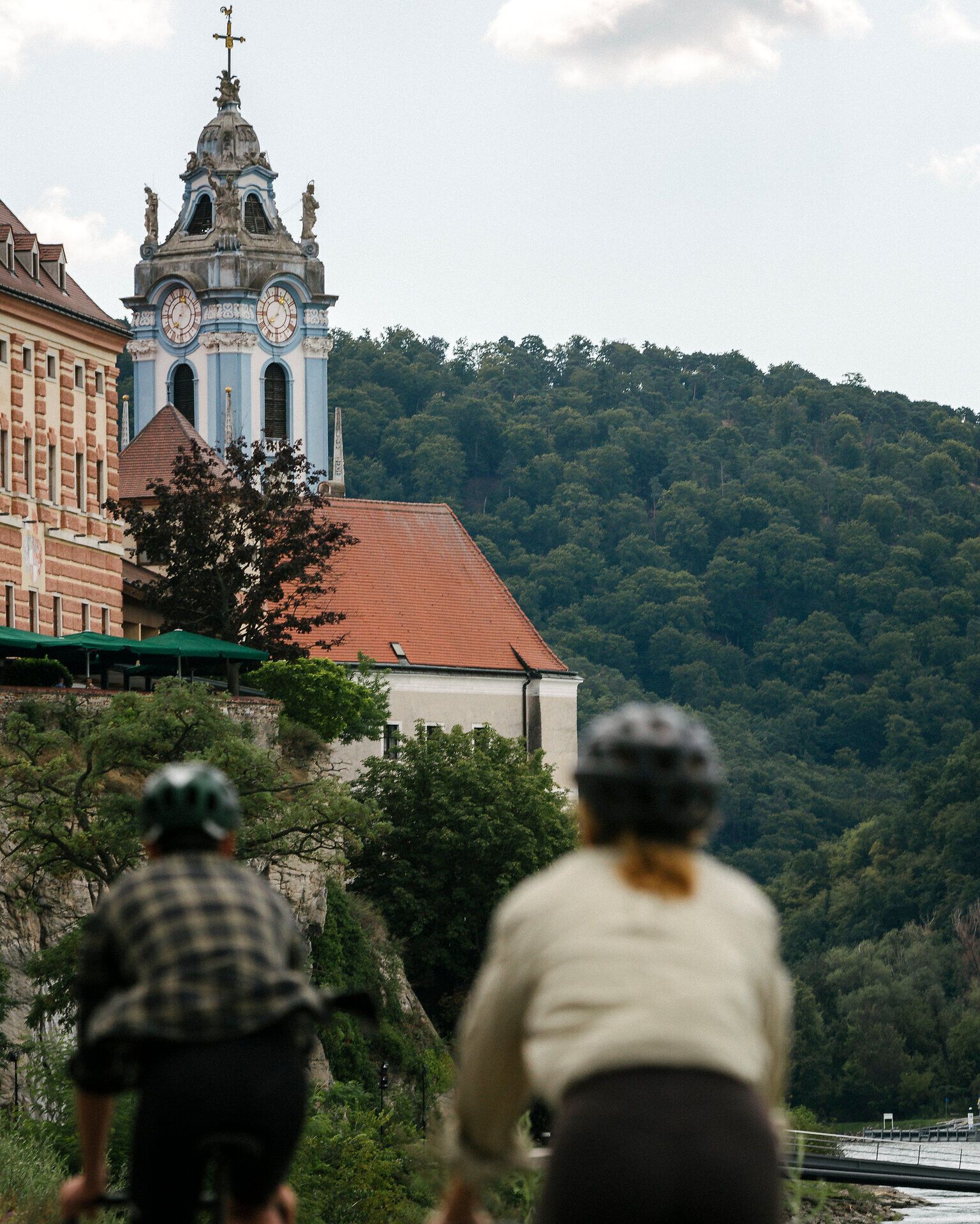 Zwei Radfahrende von hinten auf einem Weg; im Hintergrund ragt die blau-weiße Kirchturmspitze des Stifts Dürnstein über Dächer und den bewaldeten Hang.
