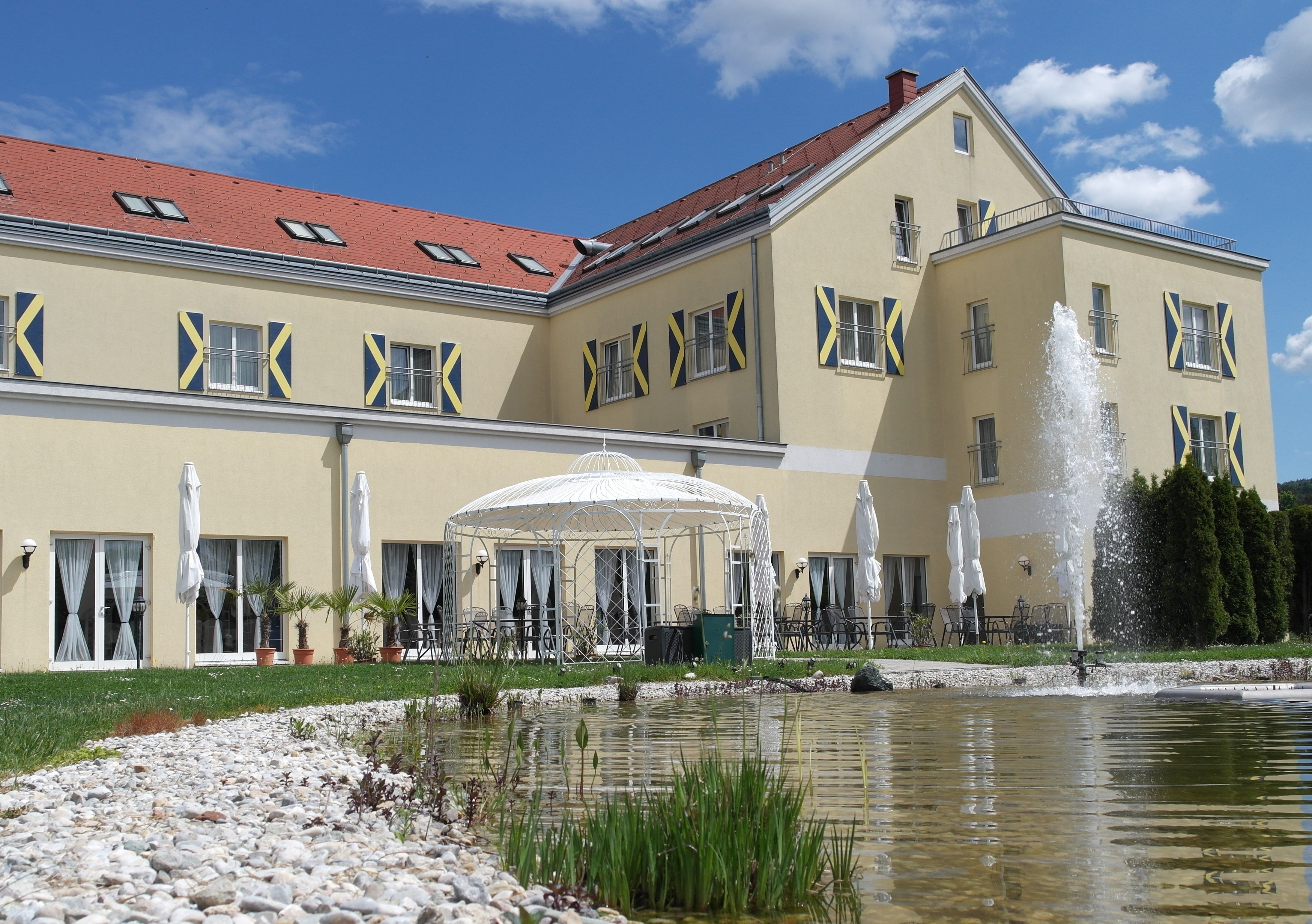 Nagy sárga épület piros tetőcseréppel, szökőkút és pavilon az előtérben.