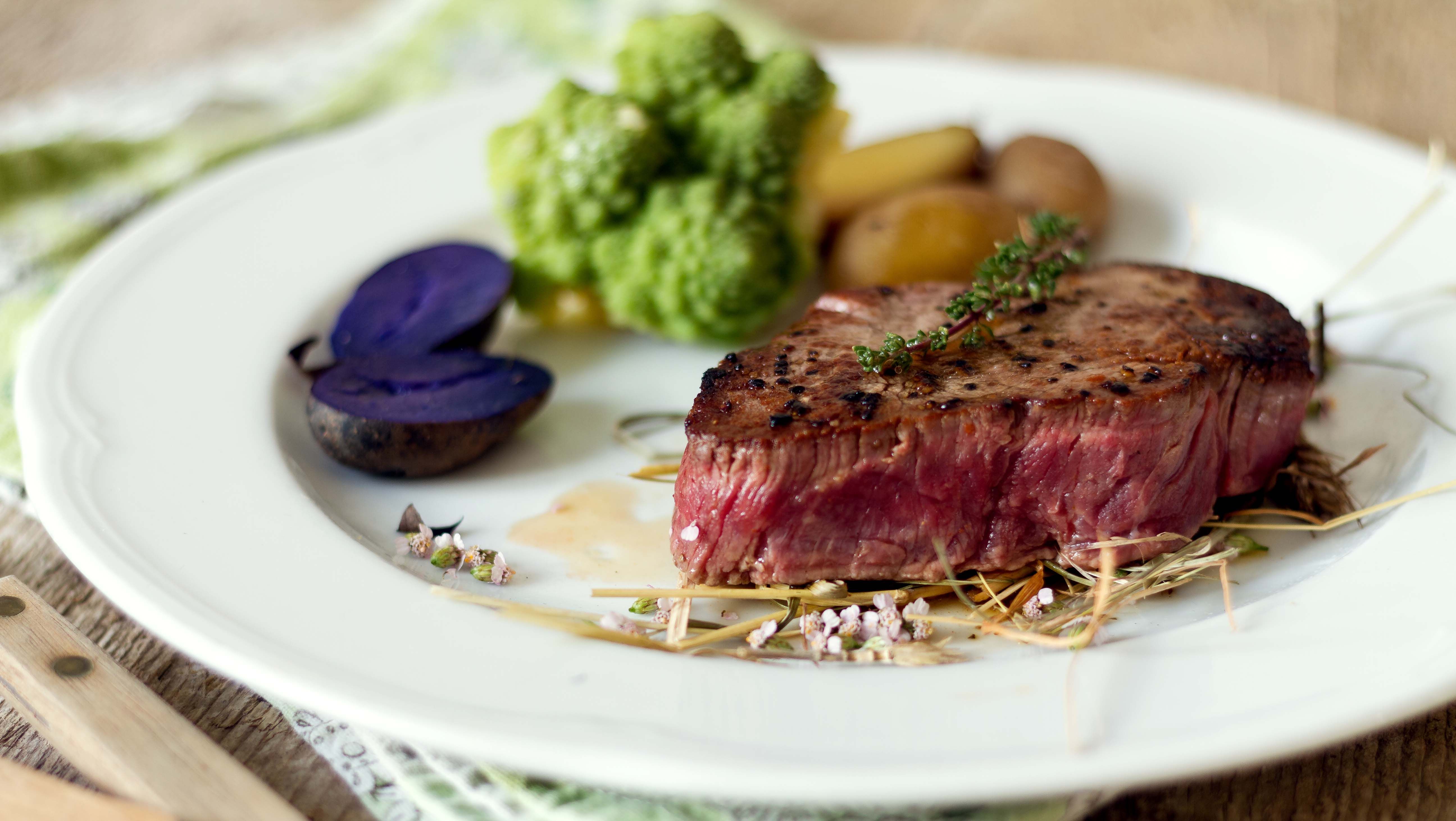 Egy szelet steak fehér kerek tányéron zöldségekkel, burgonyával és fűszernövényekkel.
