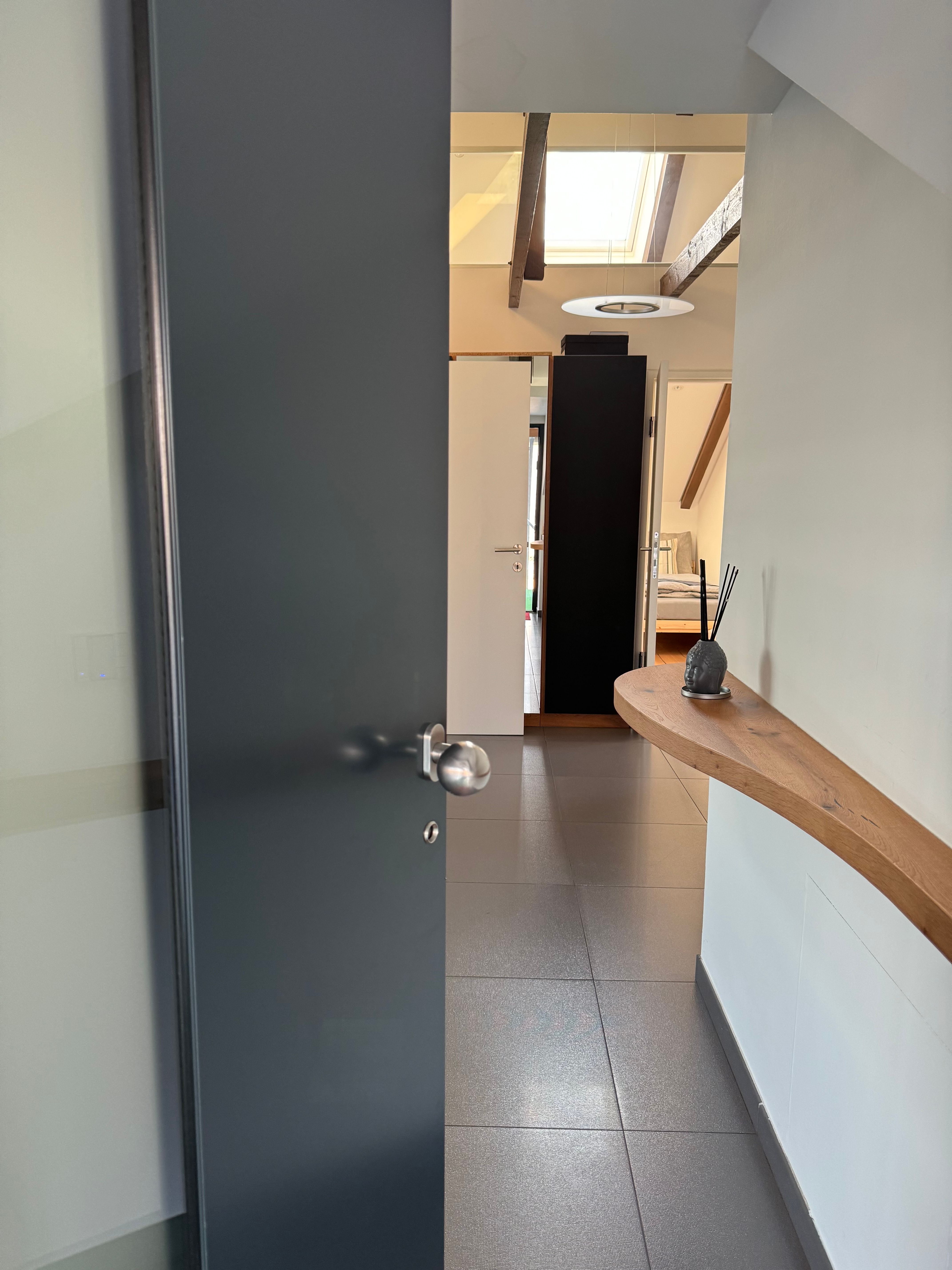 Modern bejárati rész nyitott ajtóval, csempézett padlóval és fából készült polccal a falon.