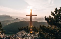 Ein majestätisches Kreuz erhebt sich vor der atemberaubenden Kulisse der sanften Hügel und Berge, während die Sonne langsam am Horizont aufgeht. Die goldenen Strahlen tauchen die Landschaft in ein warmes Licht und schaffen eine friedliche Atmosphäre, die zum Verweilen einlädt. Hier, inmitten der Natur, spürt man die Ruhe und Erhabenheit des Augenblicks.