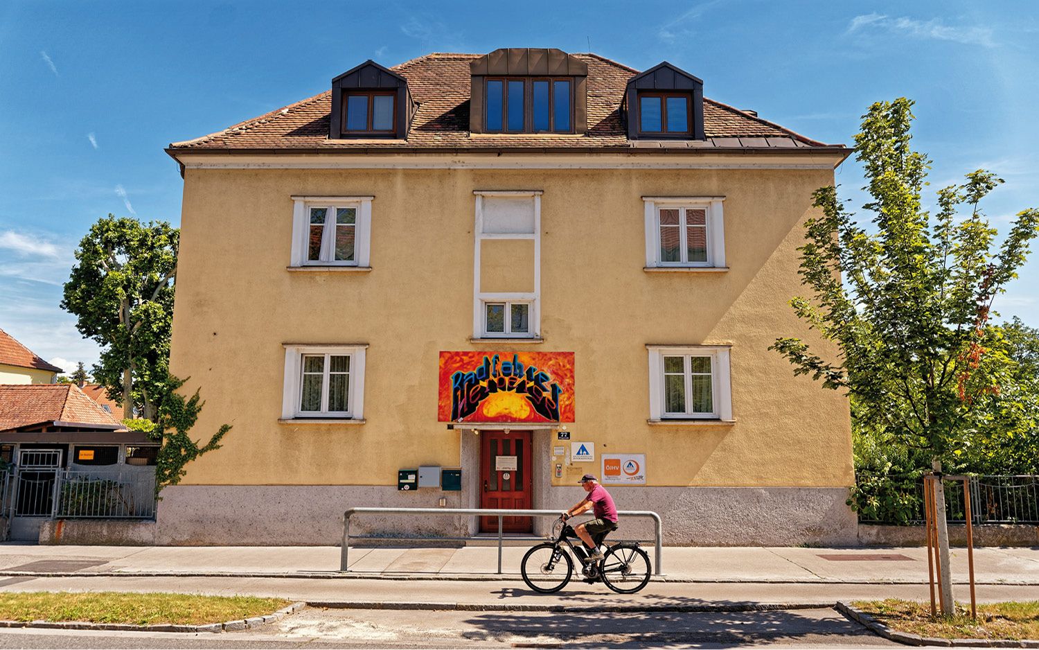 Sárga épület piros bejárattal és graffiti felirattal. Egy kerékpáros halad el mellette. Kék ég és fák a háttérben.