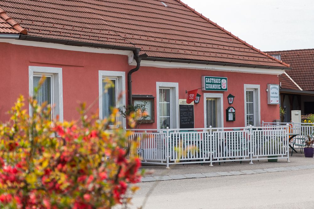 A "Gasthaus Zivanovic" feliratú rózsaszín épület, fehér kerítéssel és virágokkal körülvéve az előtérben.