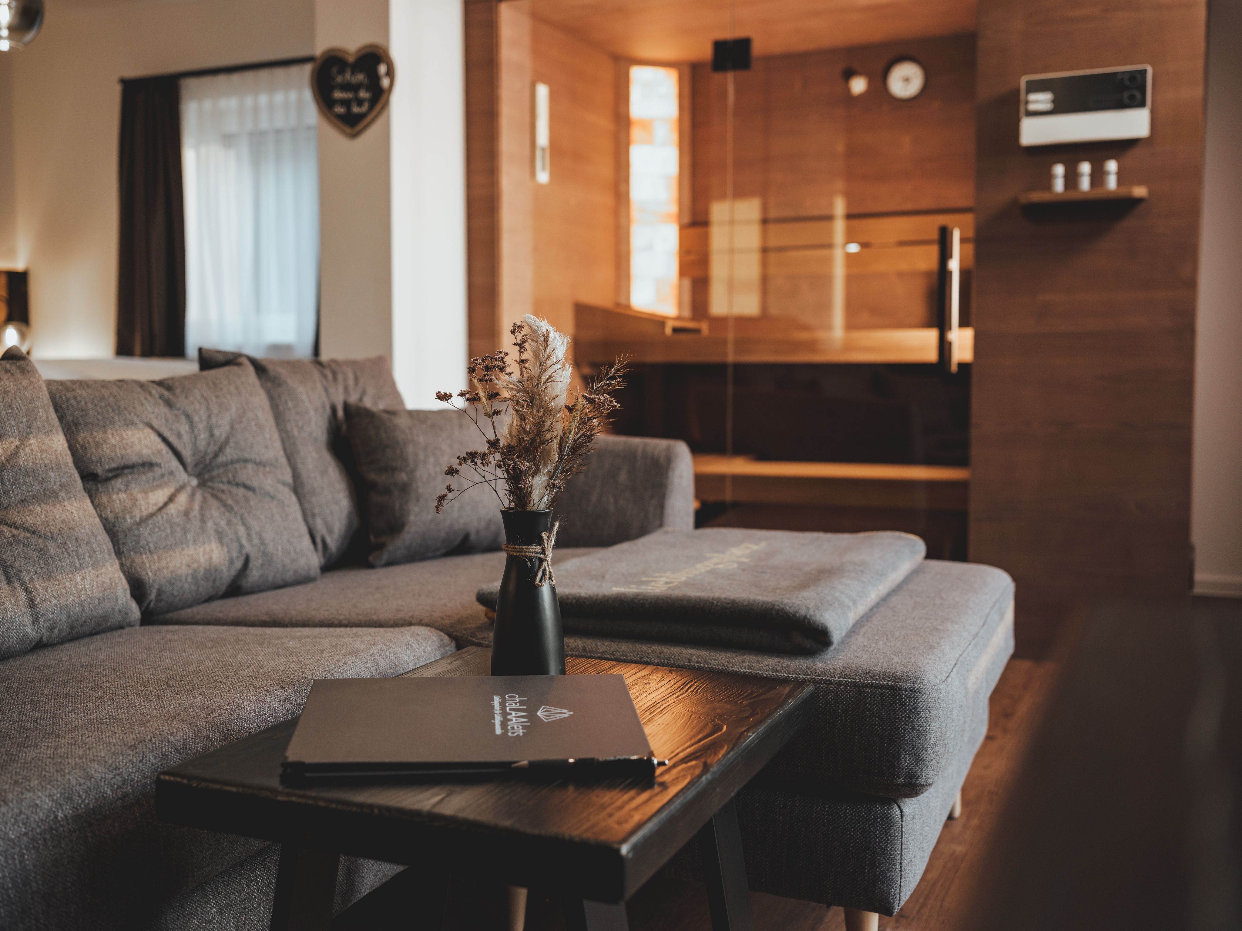 Hangulatos nappali szürke kanapéval, fa asztallal és szaunával a háttérben.