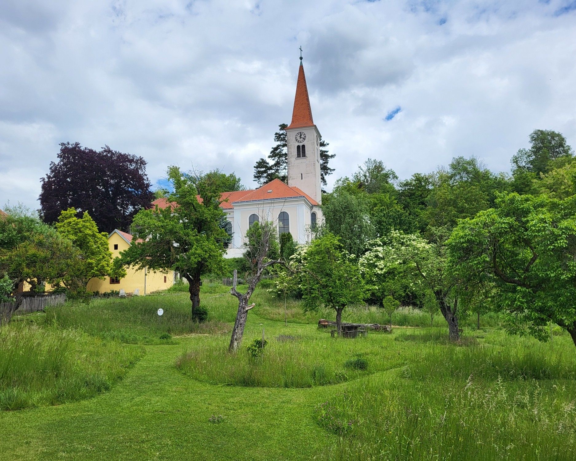 Königsbrunn am Wagramban található plébániakert, kilátással a történelmi templomra, zöld rétekkel, öreg gyümölcsfákkal és természetes kerti tájjal körülvéve.