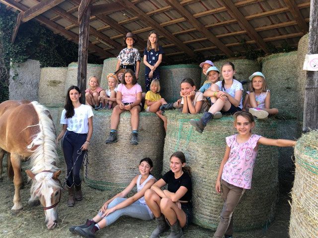 Egy csapat gyerek ül szénabálákon egy istállóban, mellettük egy ló áll. A gyerekek mosolyognak és alkalmi ruhát viselnek.