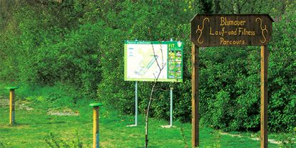Fából készült tábla "Blumauer Lauf- und Fitness Parcours" felirattal egy információs tábla előtt vidéken.