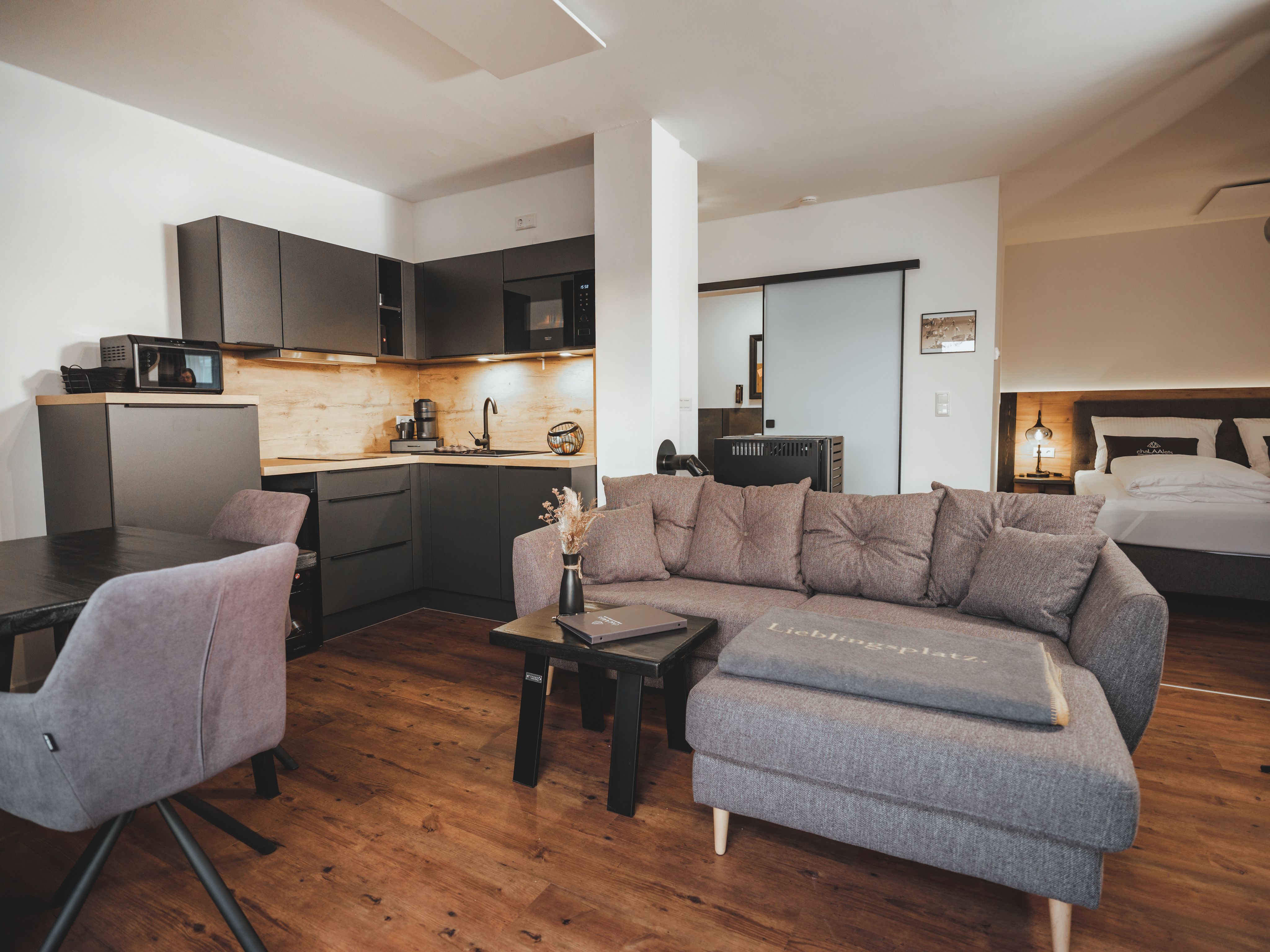 Modern nappali szürke kanapéval, konyhasarokkal és étkezőasztallal.