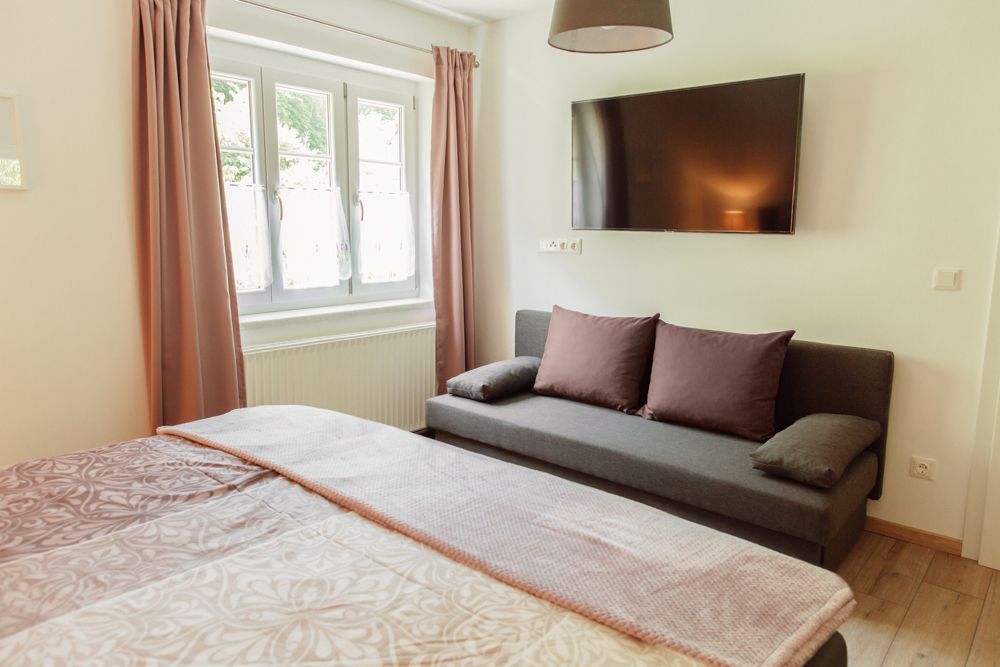 Belső nézet egy hálószobából, ágy, kanapé, ablak és falra szerelt televízió.