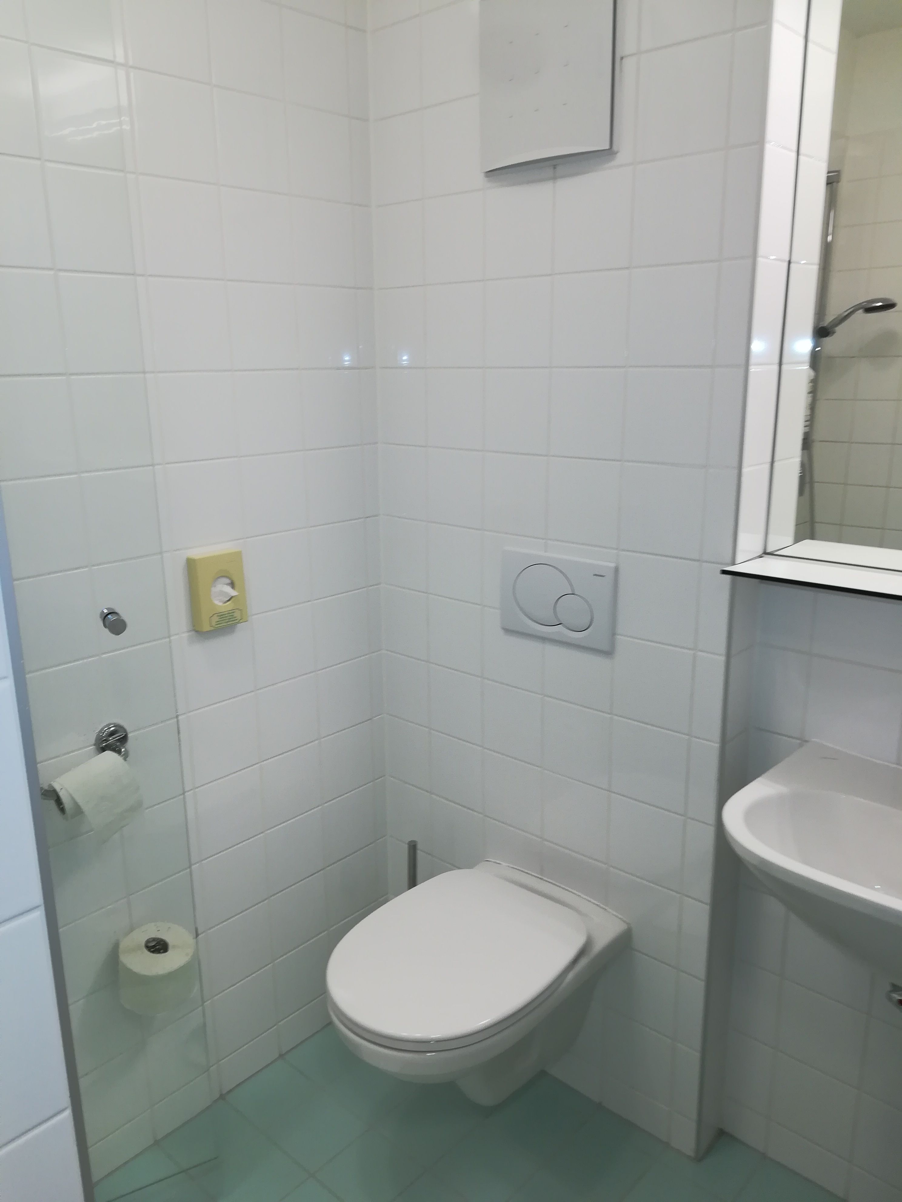 Kis fürdőszoba fehér csempézett falakkal, WC-vel, mosdókagylóval és WC-gurigatartóval.