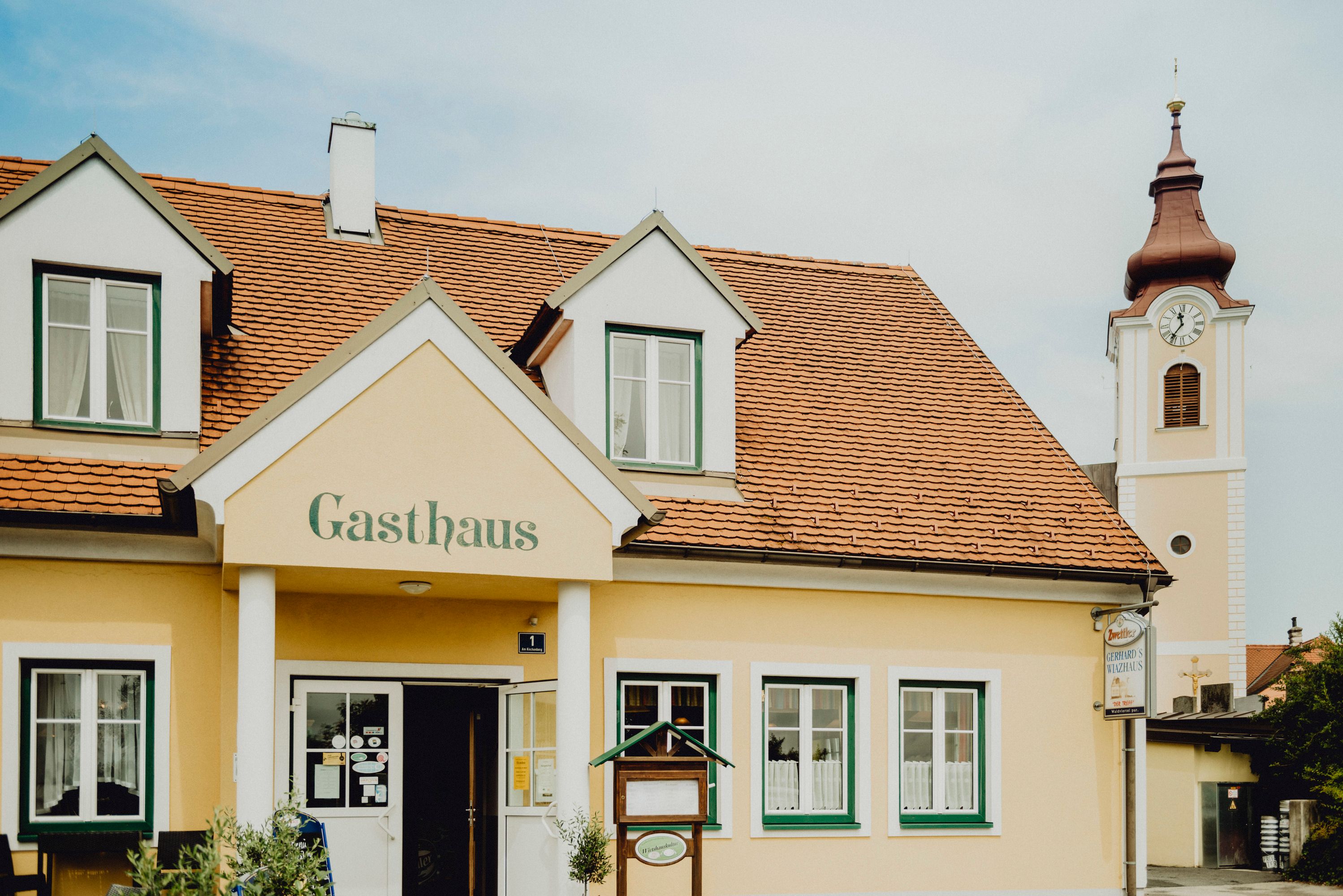 Sárga színű fogadó piros tetőcseréppel és a háttérben egy templomtoronnyal.