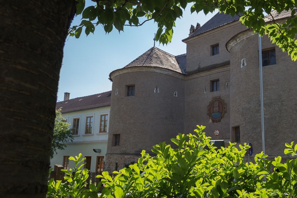 Történelmi épület kerek tornyokkal és címerrel, zöld fákkal és bokrokkal körülvéve.