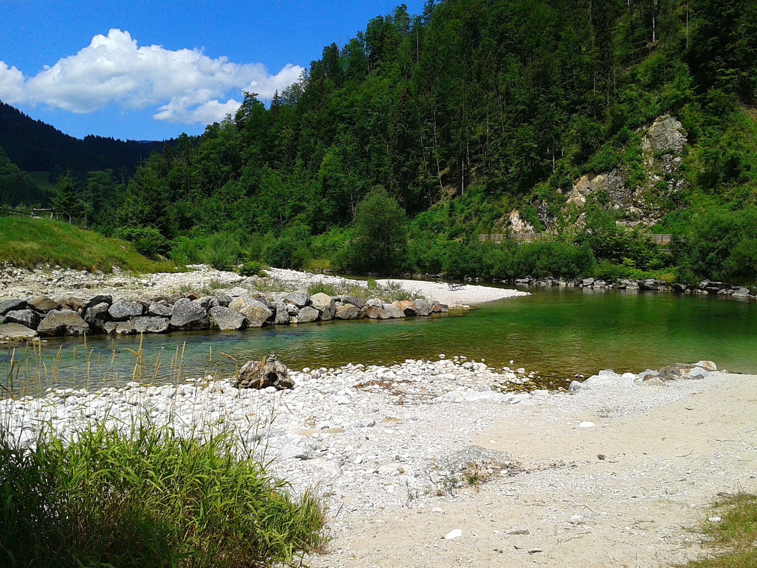 Folyami táj tiszta vízzel, kavicsos parttal és erdős dombokkal.