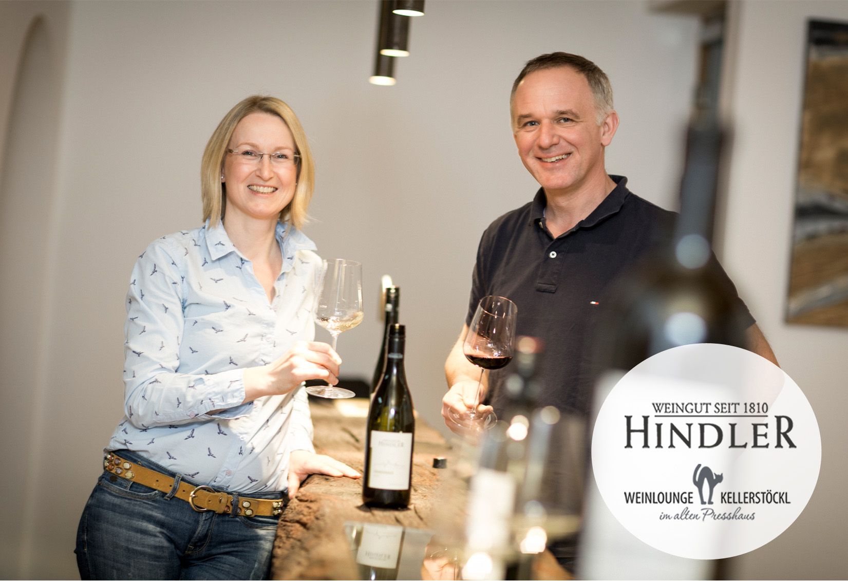 Egy borászpáros áll mosolyogva, borospoharakkal a kezükben a Hindler pincészet egyik asztalánál.