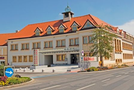 Traiskircheni városháza piros tetővel és több ablakkal, utcával és virágágyásokkal körülvéve.