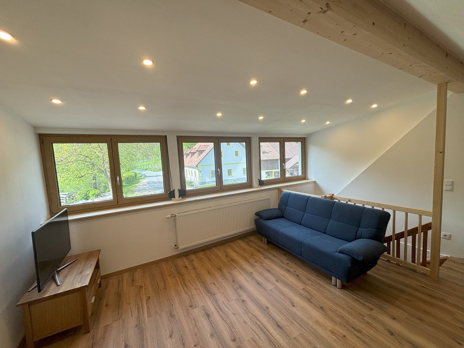 Nappali kék kanapéval, fapadlóval és nagy ablakokkal, kilátással a szabadba.