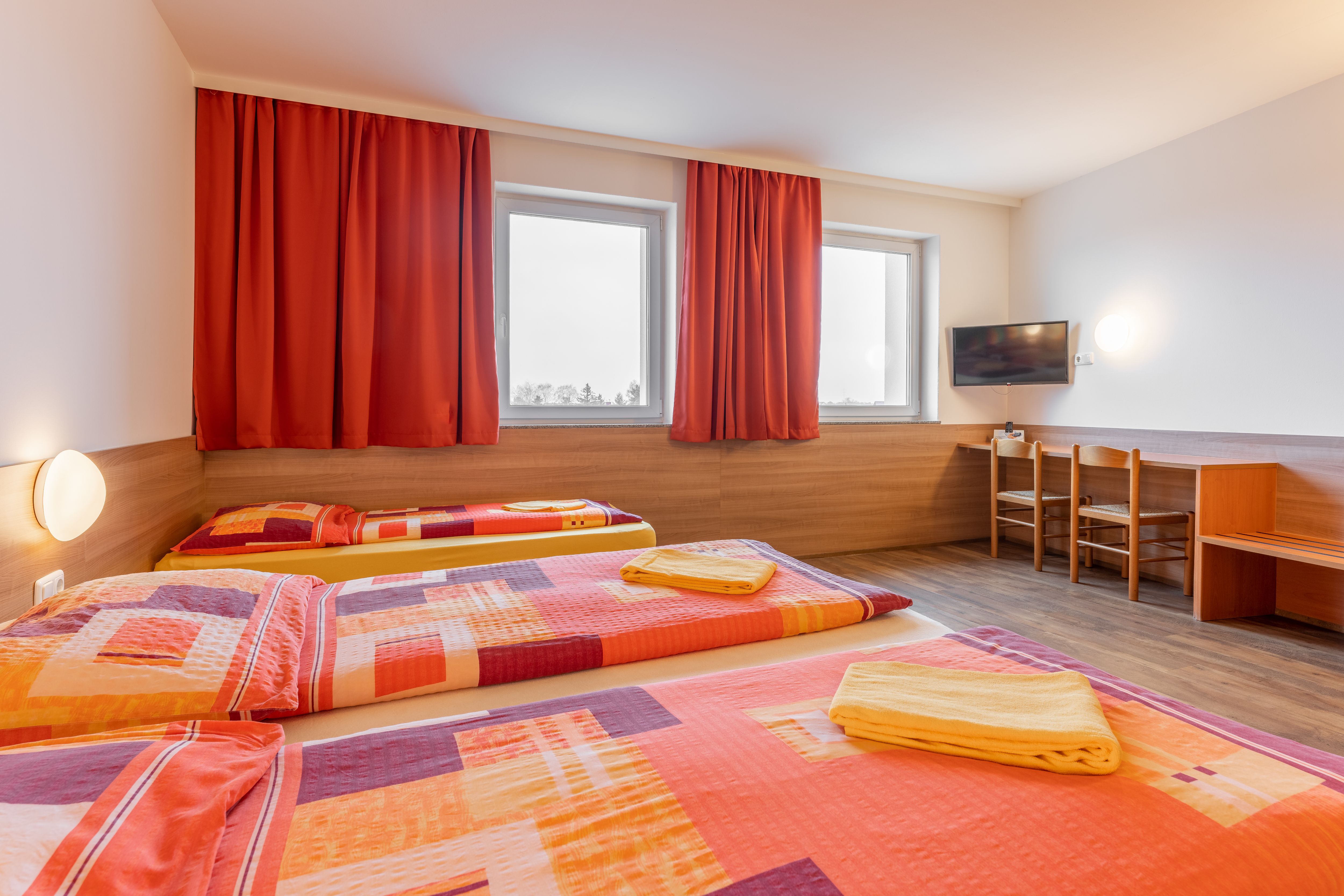 Hotelszoba három ággyal, piros függönyökkel és egy TV-vel a falon.