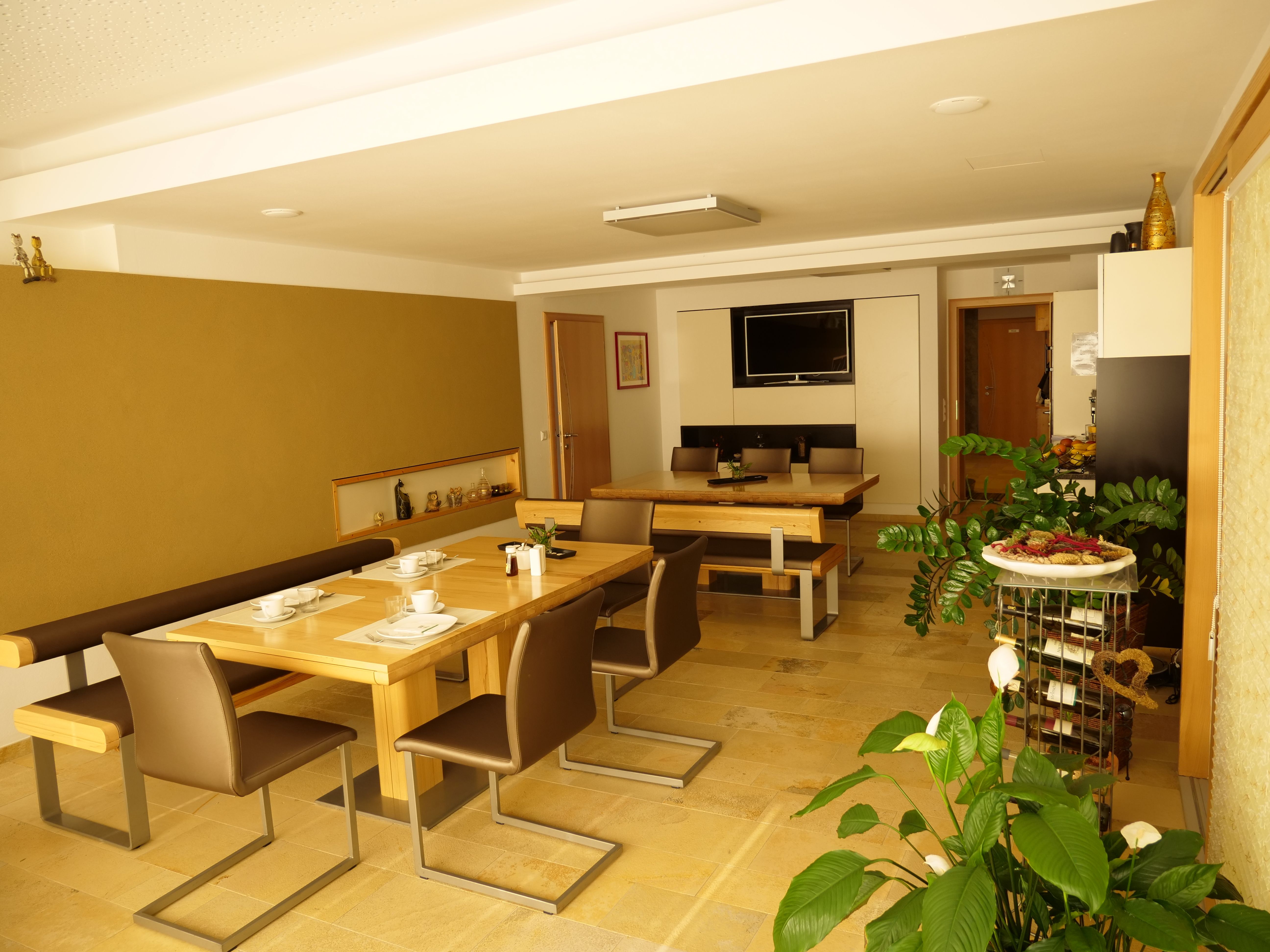 A reggelizőterem faasztalokkal, székekkel és növényekkel, edényekkel díszített, a falon TV-vel.