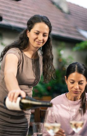 Eine Winzerin schenkt im Gastgarten des Weinguts Herzog in Bad Vöslau Wein für ihre Gäste ein – Heurigenkultur und Jause in entspannter Atmosphäre im Wienerwald in Niederösterreich.