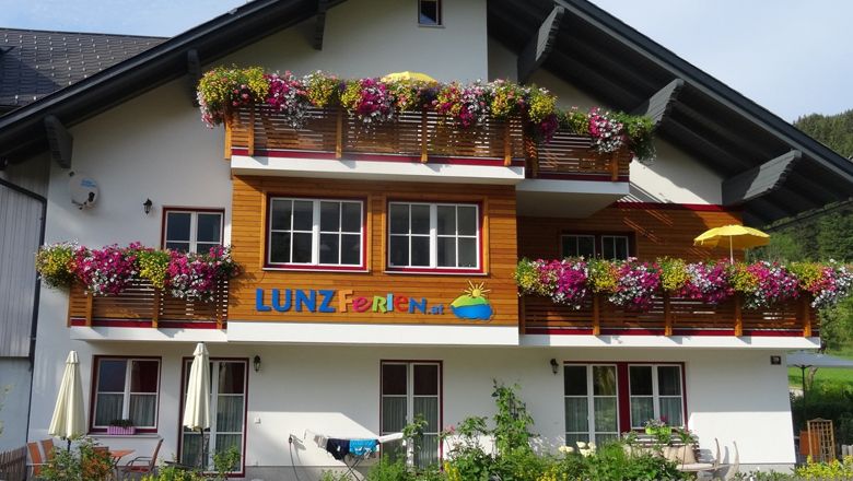 Nyaralóház színes virágokkal az erkélyeken és a "LUNZ Ferien" felirattal.