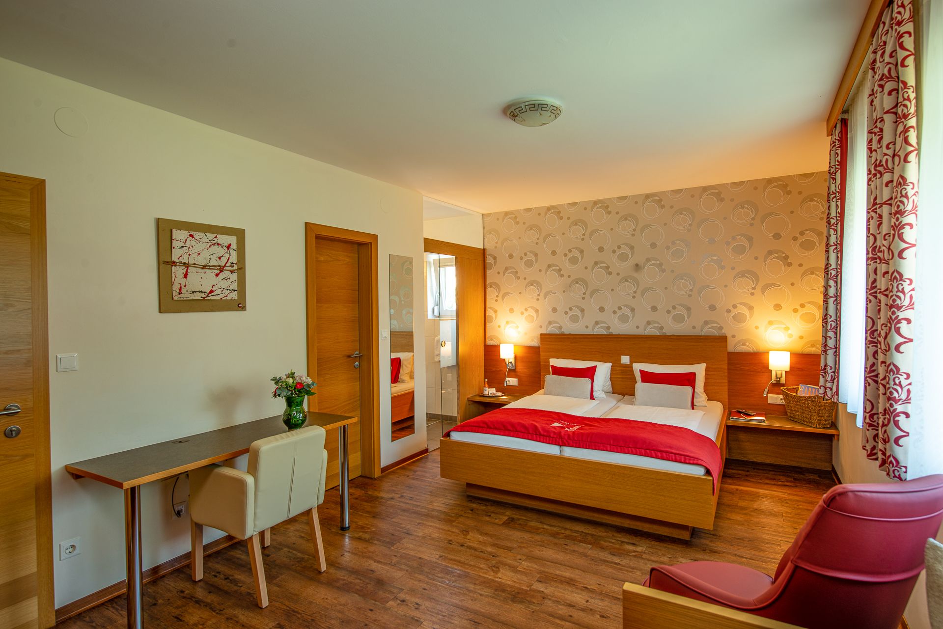 Hotelszoba franciaággyal, íróasztallal, székkel és piros karosszékkel.