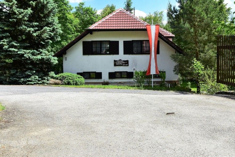 Fehér ház, piros tetővel, előtte osztrák zászlóval, fákkal körülvéve.