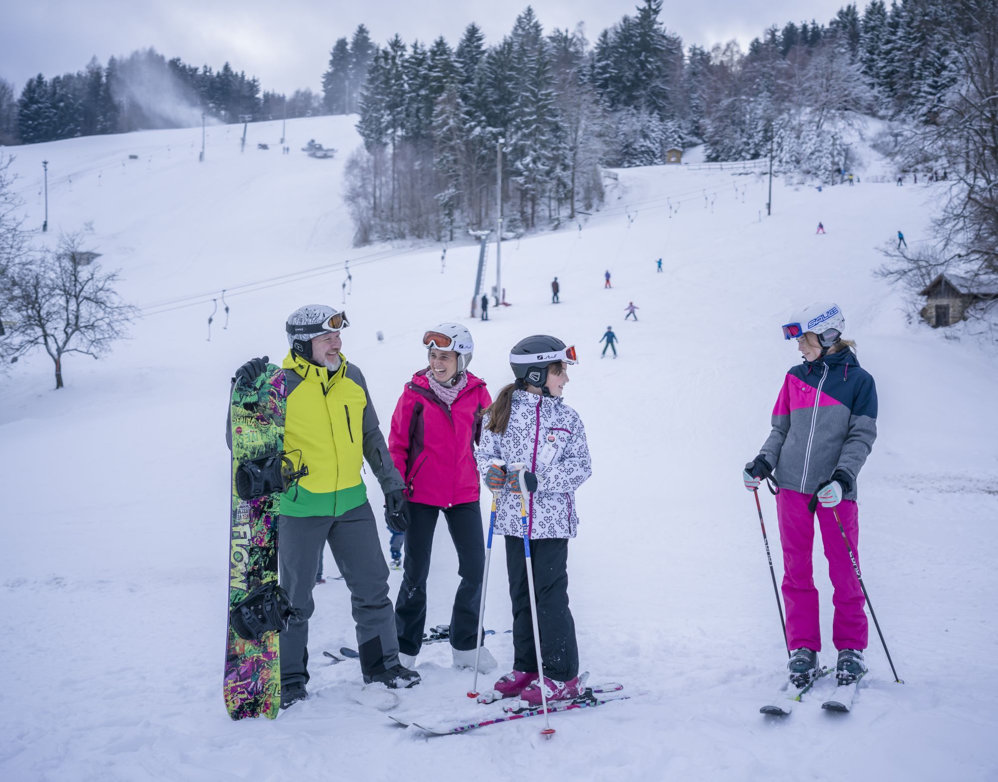 Síelők és snowboardosok csoportja a hóban egy felvonóval ellátott sípálya előtt.