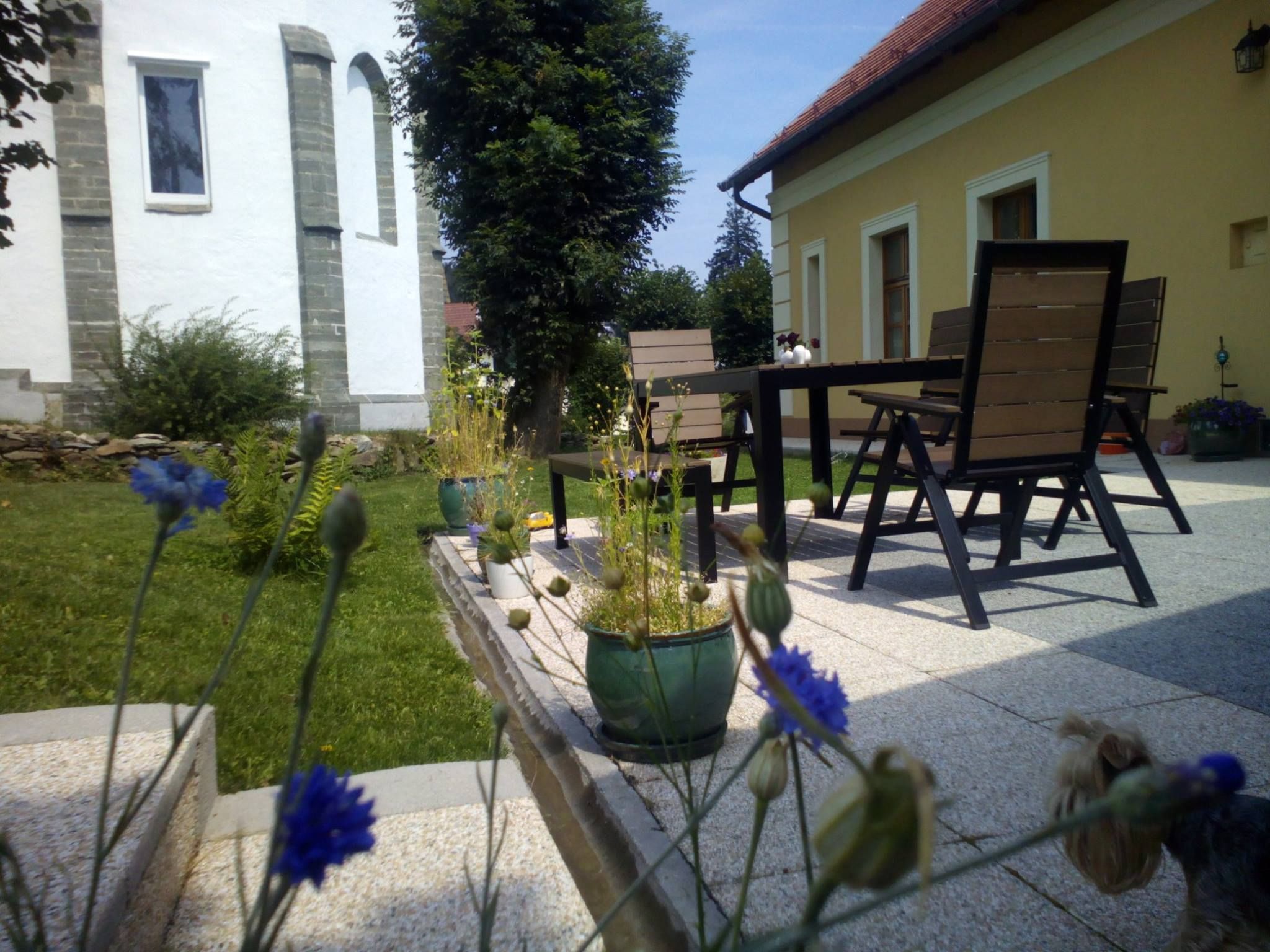 Terasz asztallal, székekkel és növényekkel egy sárga ház melletti kertben.