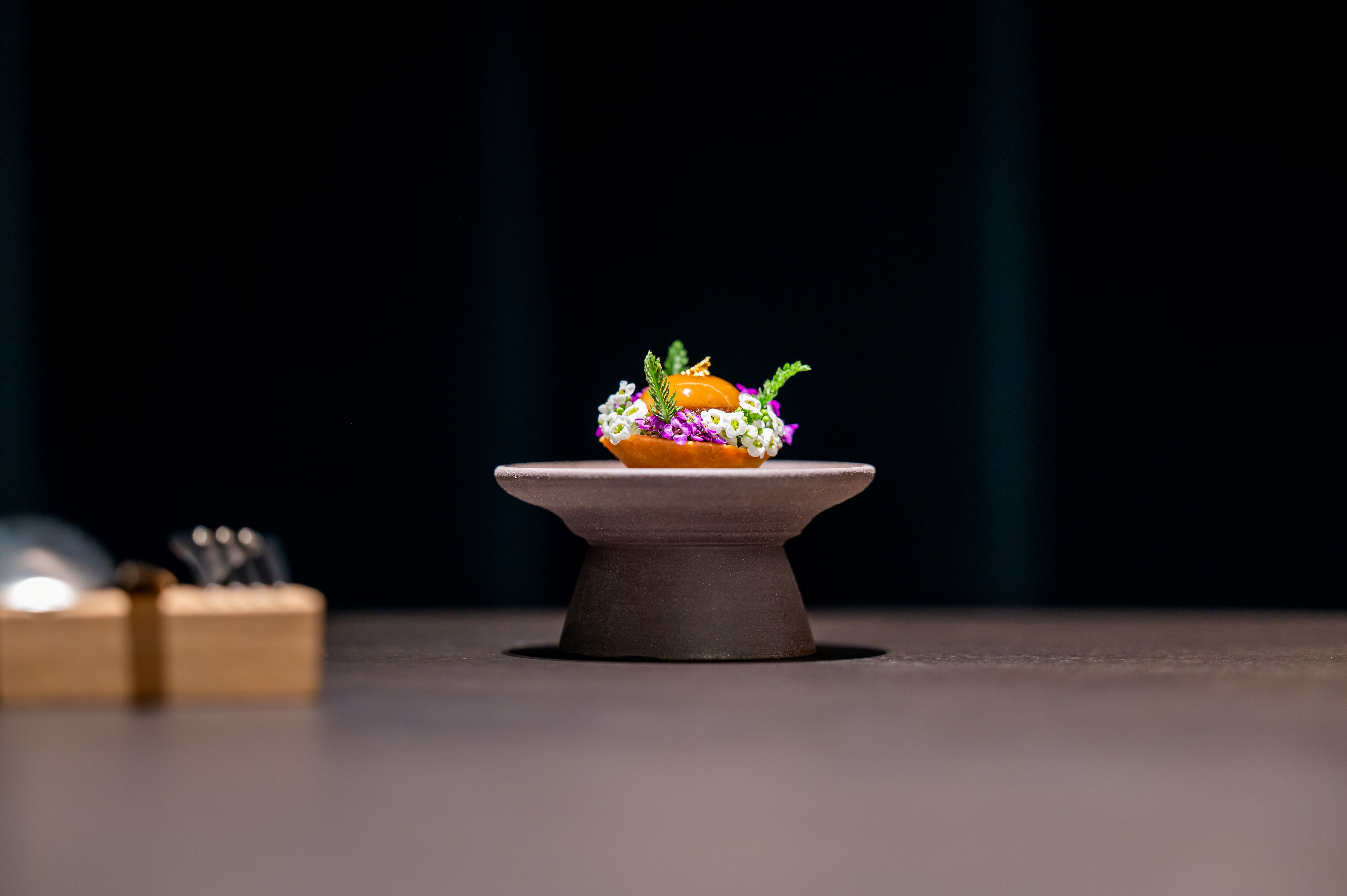 Művészien elrendezett tál egy kis talapzaton, virágokkal és gyógynövényekkel körülvéve, sötét háttér előtt.