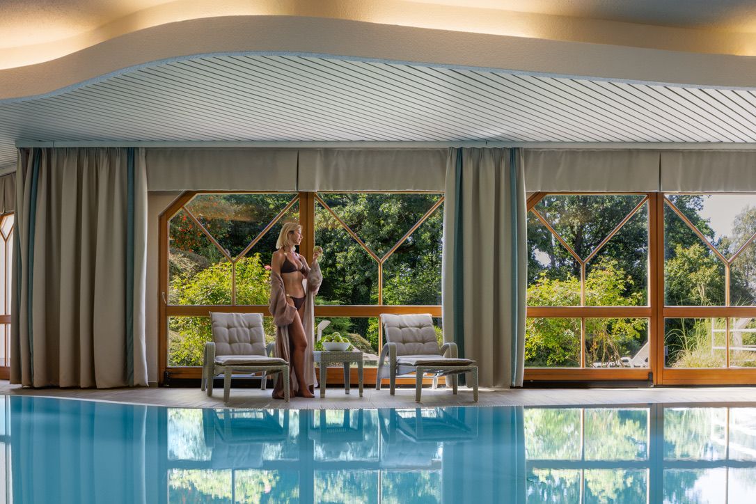 Egy fürdőruhás nő áll egy fedett medence mellett, amelynek nagy ablakai zöld fákra néznek.
