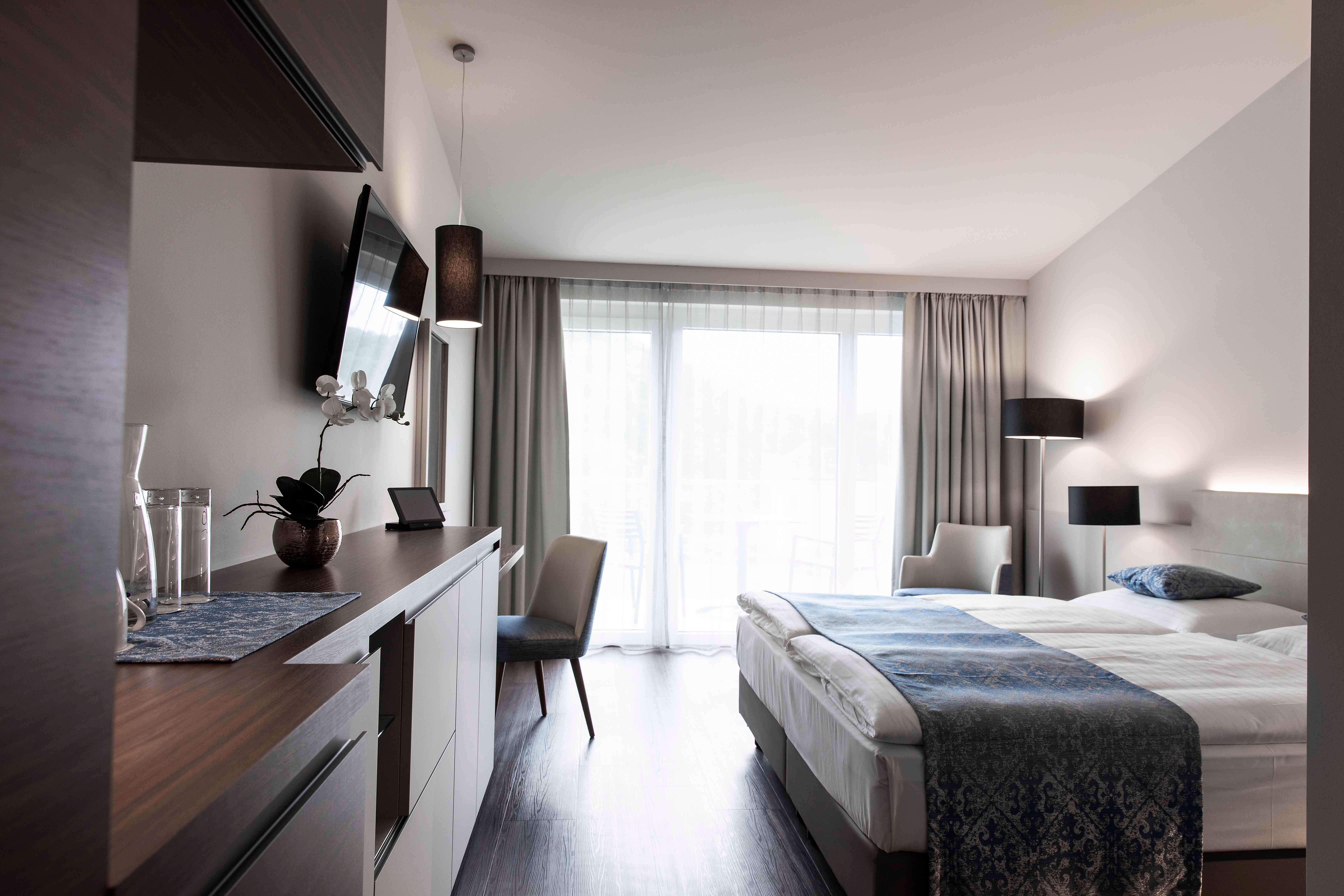 Modern szállodai szoba franciaággyal, TV-vel, íróasztallal és nagy, függönyös ablakokkal.