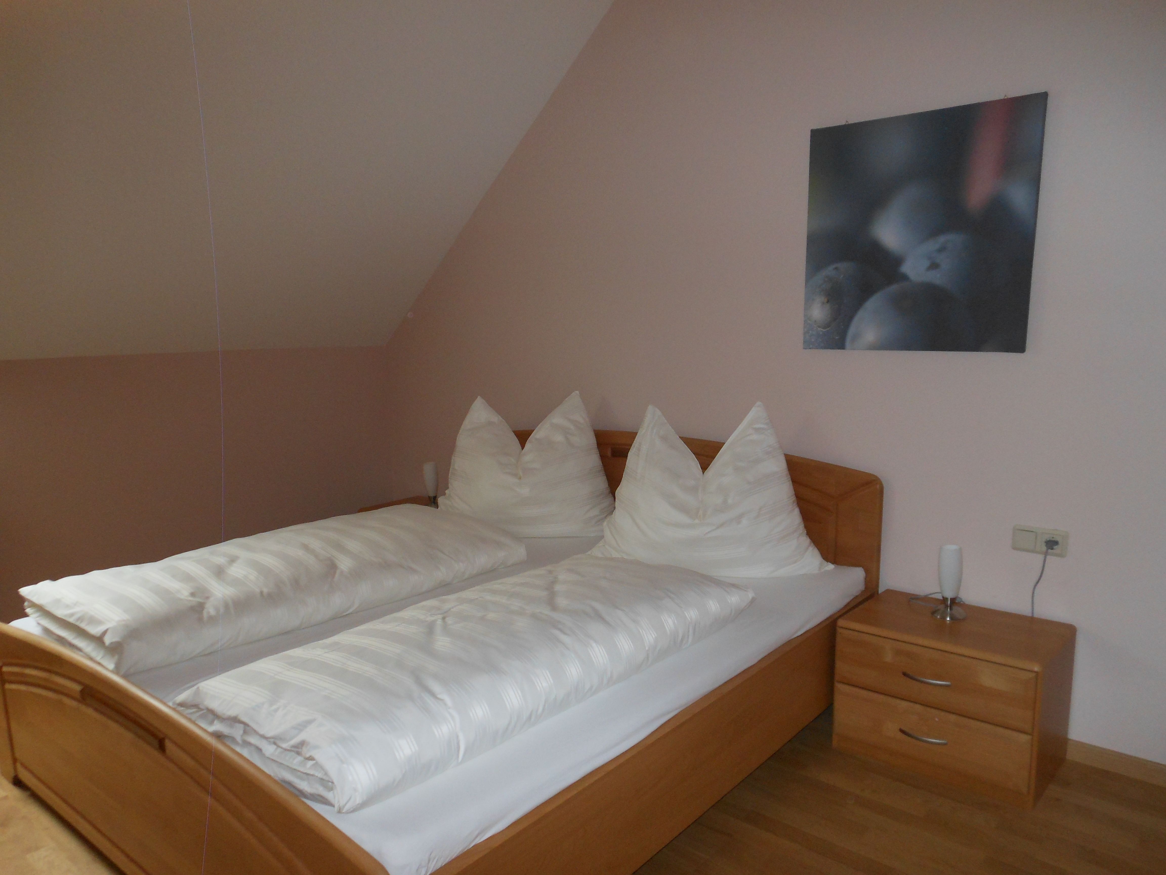 Egyszerű vendégszoba franciaággyal, fehér paplannal és éjjeliszekrénnyel, lámpával.