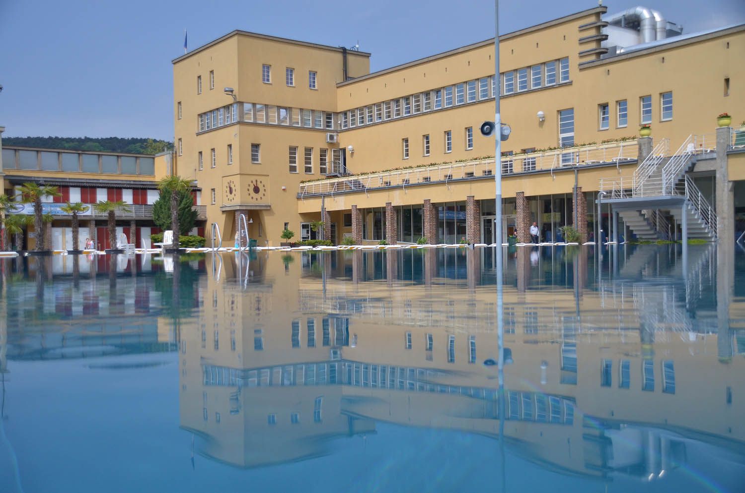 Egy szabadtéri medence, a háttérben egy nagy sárga épület, amely tükröződik a vízben.