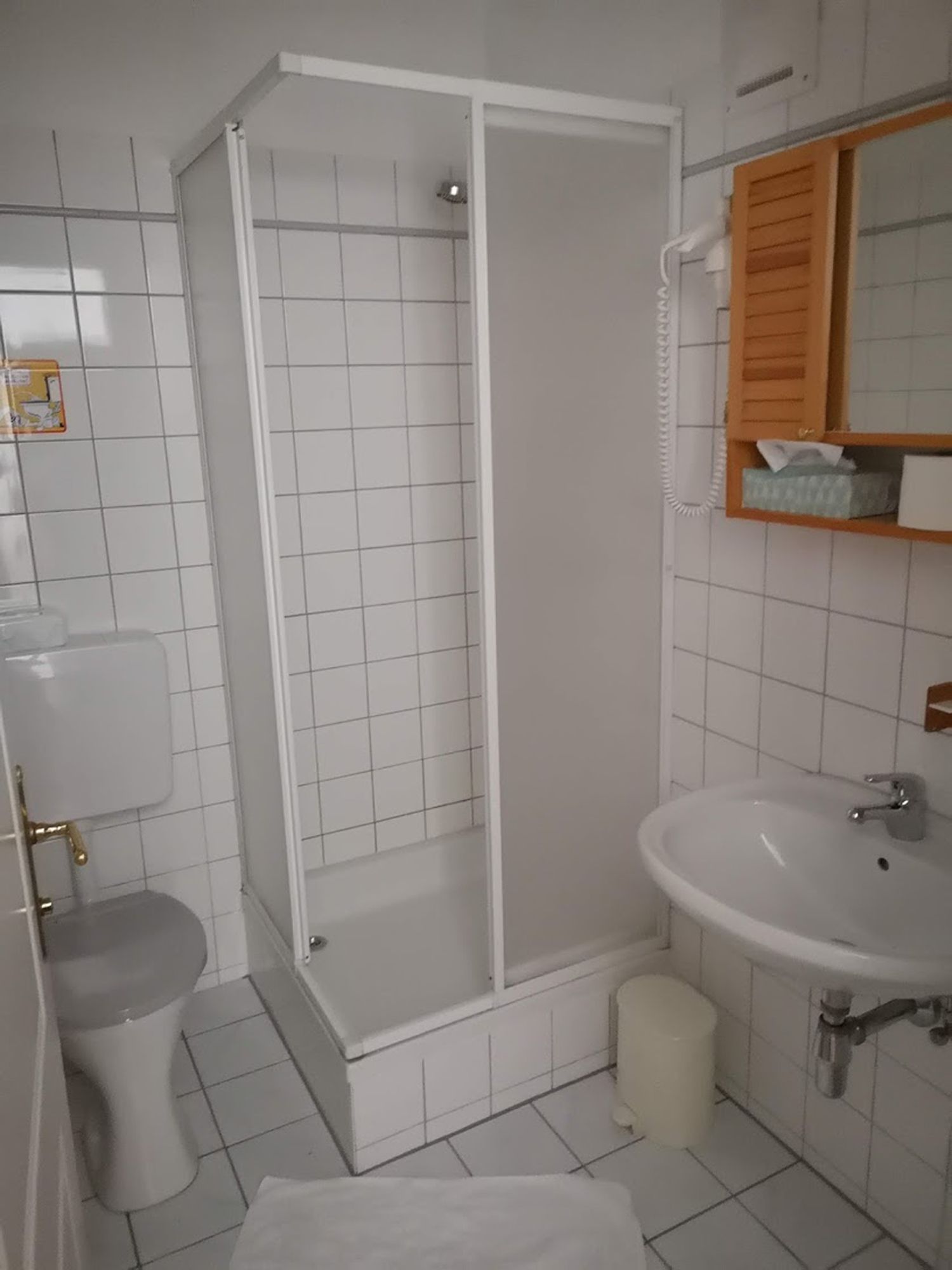 Egy kis, fehér csempézett fürdőszoba zuhanyzóval, WC-vel és mosdókagylóval.