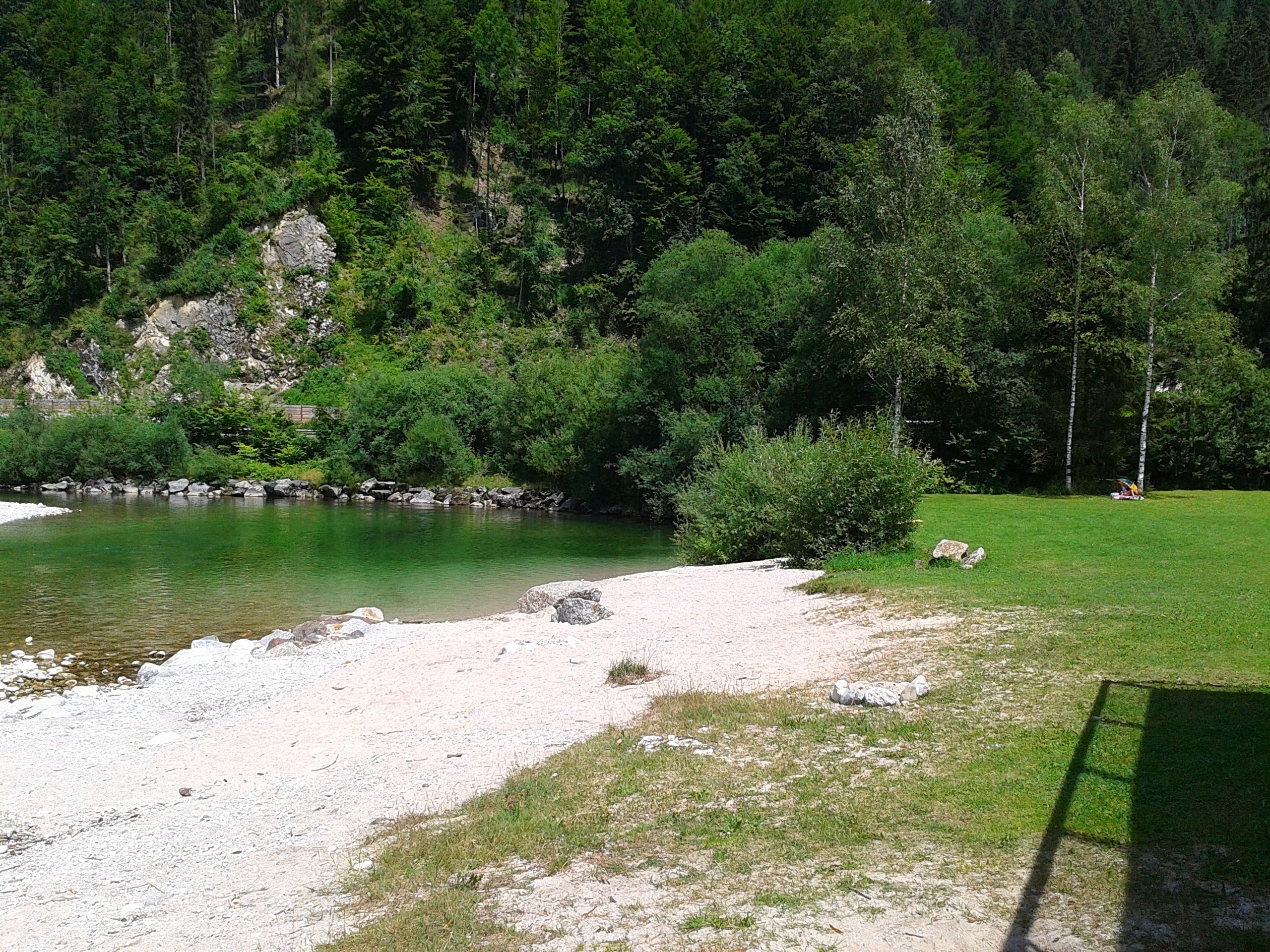 Tiszta vizű folyó, zöld fákkal körülvéve, a parton kis kavicsos terület.