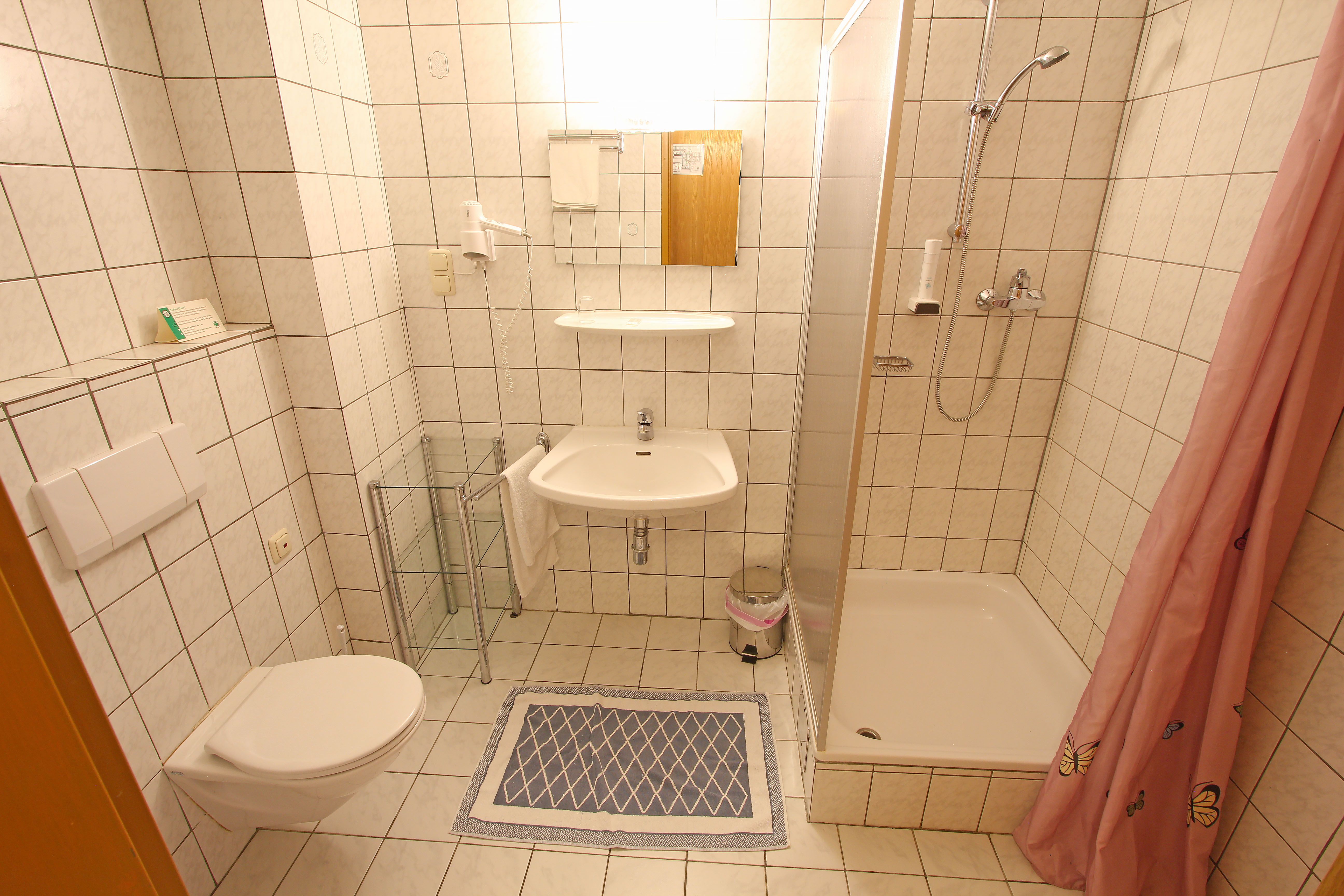 Kis fürdőszoba zuhanyzóval, mosdóval és WC-vel, fehér csempével.