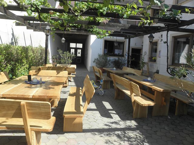 Fából készült terasz asztalokkal és padokkal egy szőlővel borított pergola alatt.