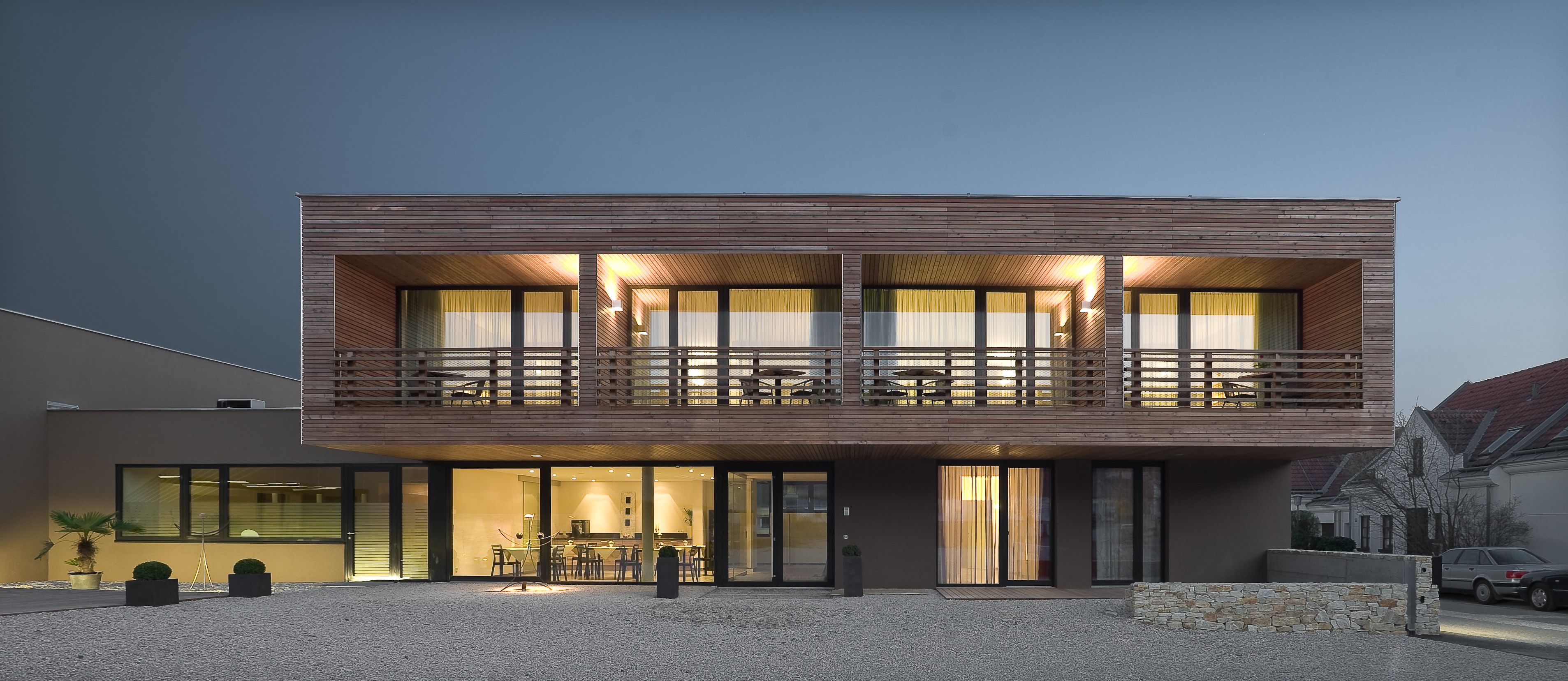 Modern kétszintes ház, fa burkolattal és nagy ablakokkal, éjszakai világítással.