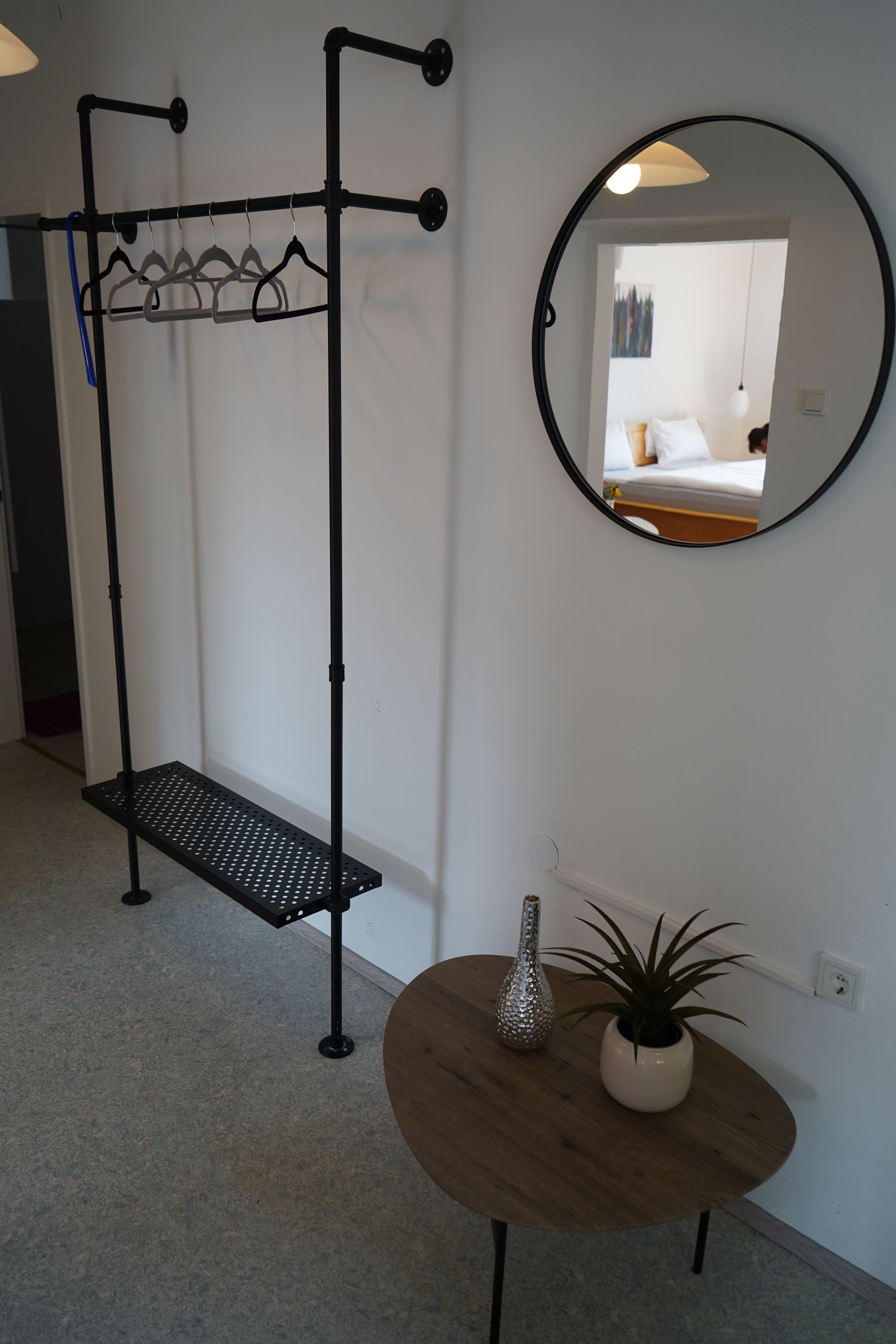 Belső nézet egy szobából, ruhatartóval, kerek tükörrel és kis asztallal, növénnyel és vázával.