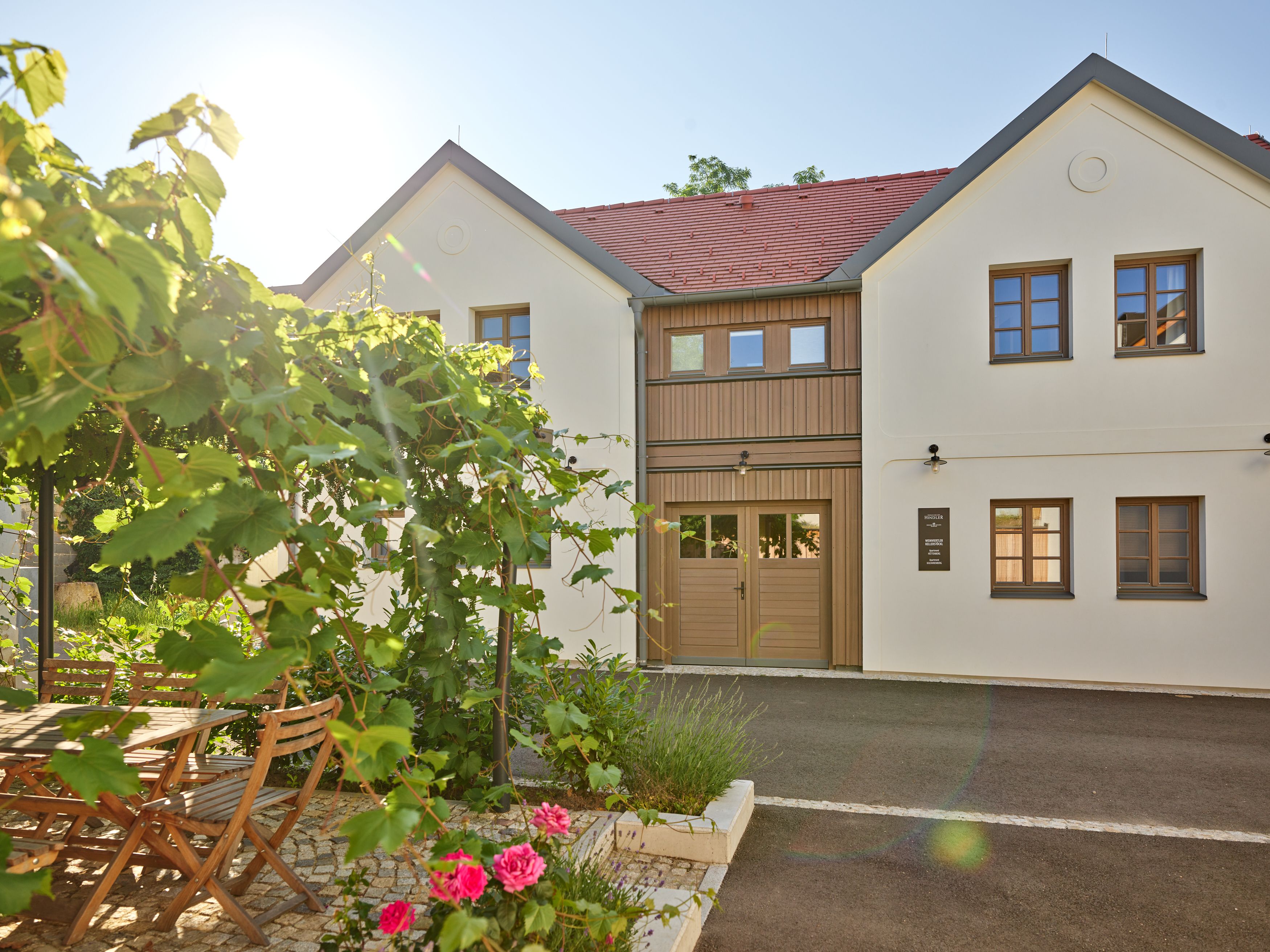 Modern, kétszintes épület, fehér falakkal és piros tetőcseréppel. Az előtérben kert, faasztallal, székekkel és virágos növényekkel.