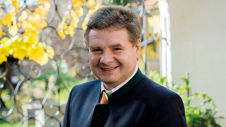 Egy mosolygó, hagyományos ruhás férfi áll kint egy kovácsoltvas kerítés előtt, a háttérben sárga levelekkel.