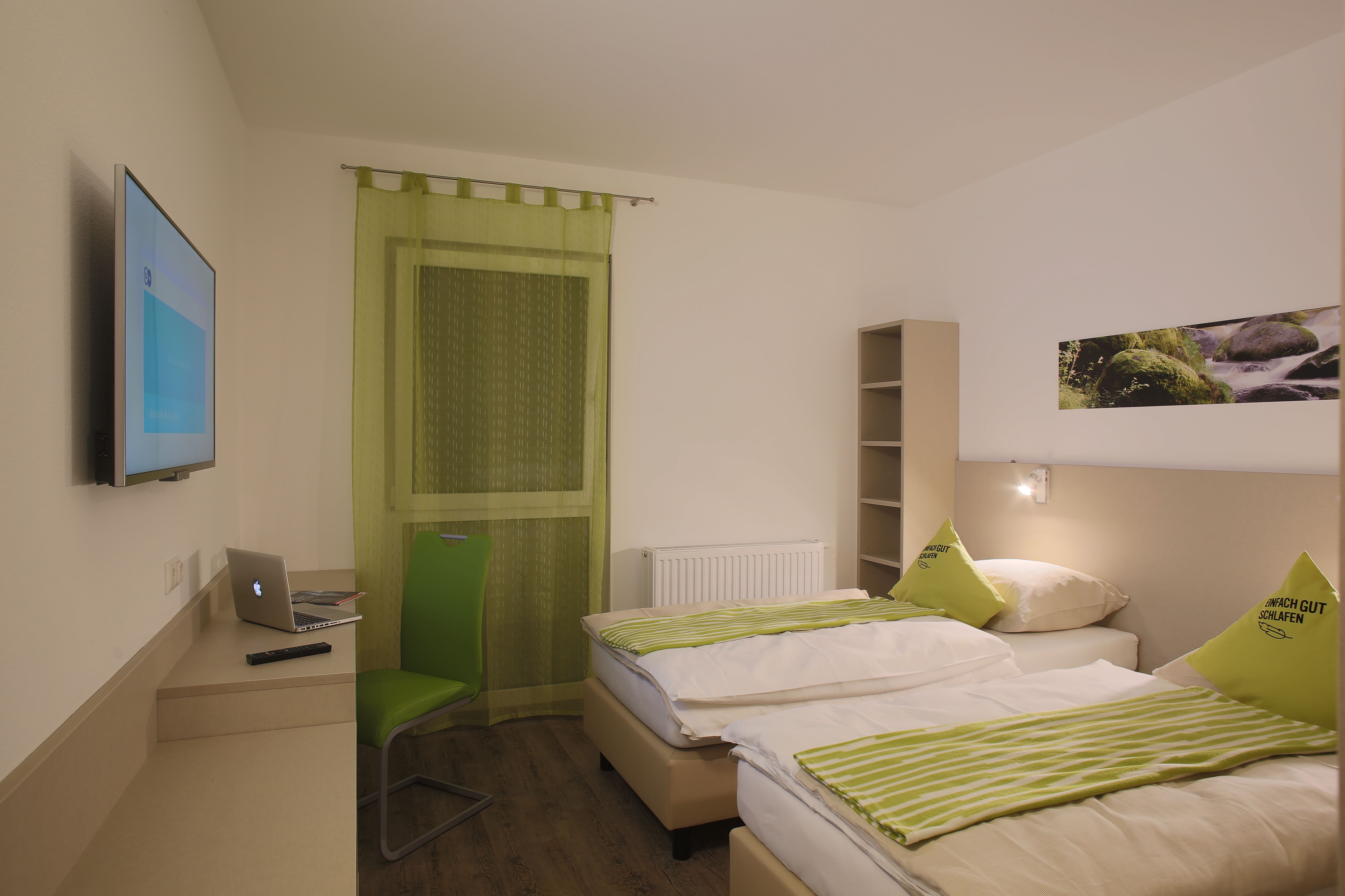 Modern motelszoba két ággyal, zöld székkel, asztalon lévő laptoppal és falra szerelt TV-vel.