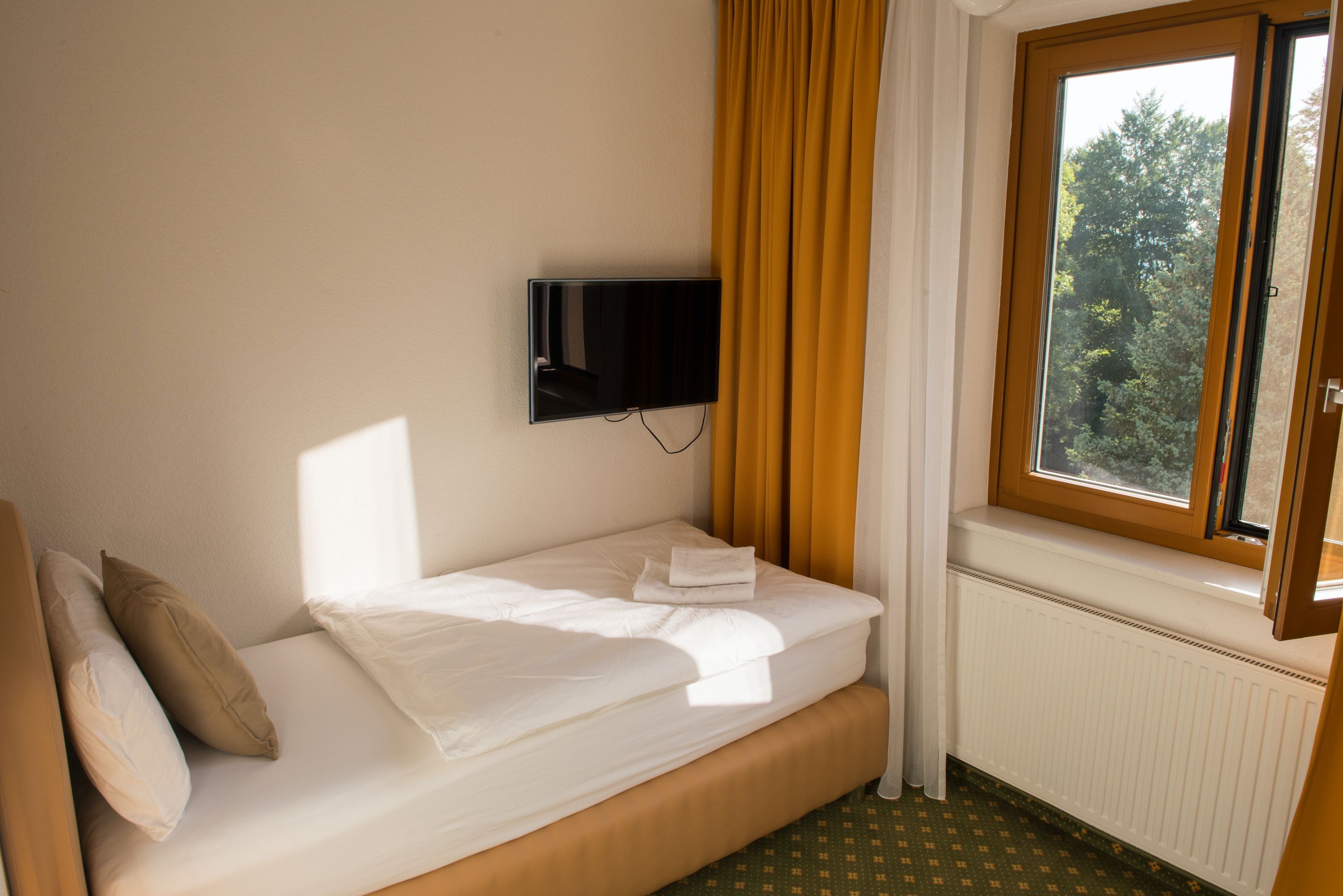 Egyágyas szoba egyszemélyes ággyal, sárga függönyökkel, zöldre néző ablakkal és falra szerelt TV-vel.