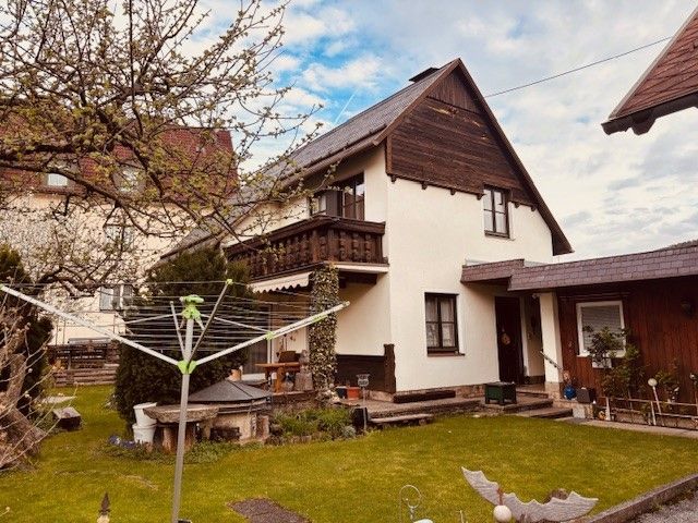 Egy kétszintes ház erkéllyel és kerttel, fákkal körülvéve, előtérben egy szárítógép.