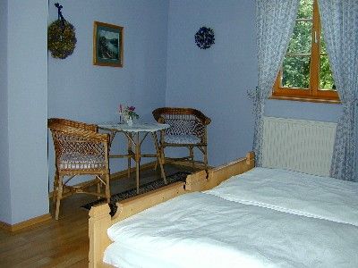 Hangulatos szoba két ággyal, kis asztallal és két székkel. Az ablakokon függönyök és fali díszek láthatók.