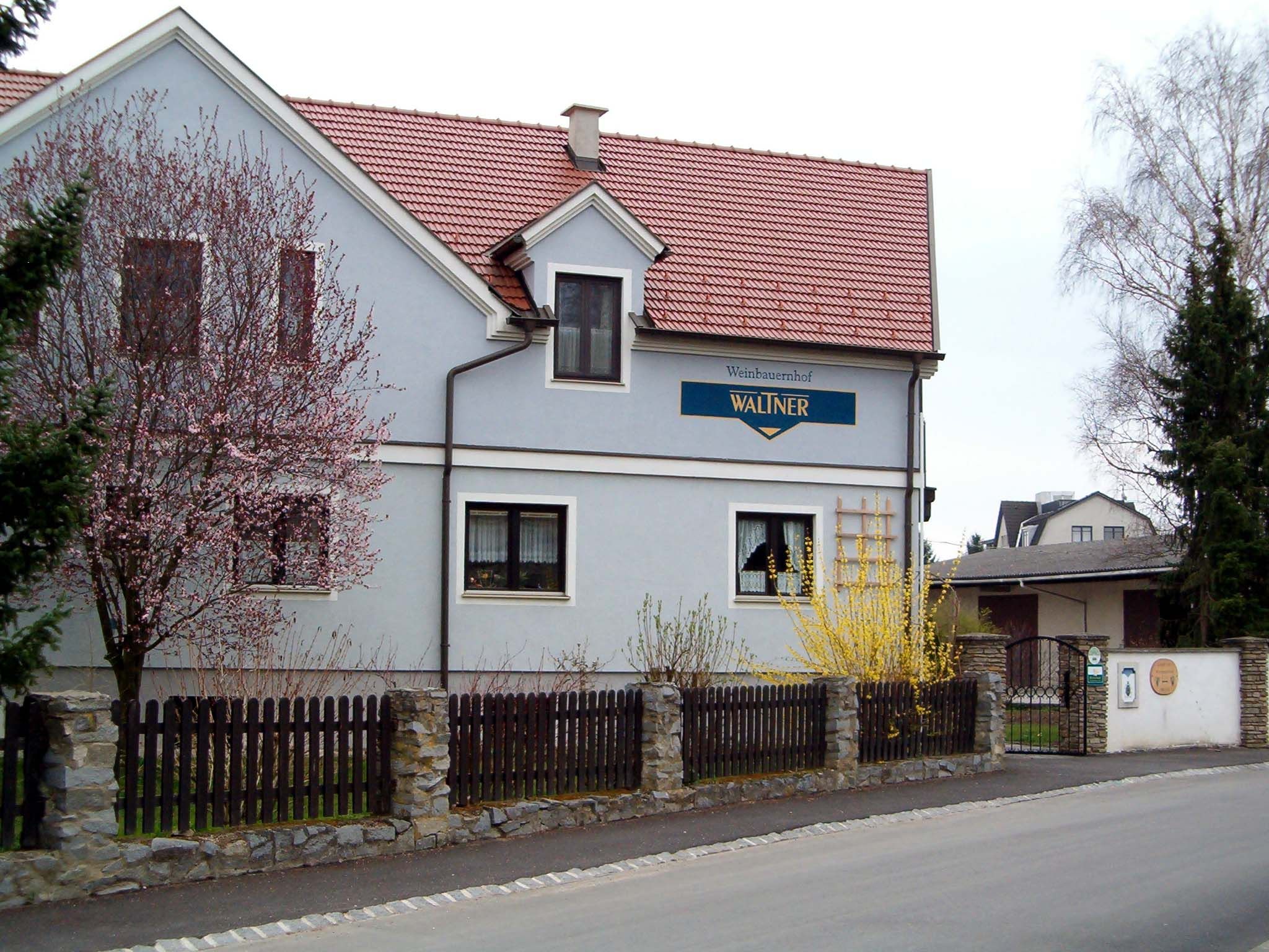 Egy kétszintes ház piros tetővel és kék homlokzattal, virágzó fákkal és fakerítéssel körülvéve.