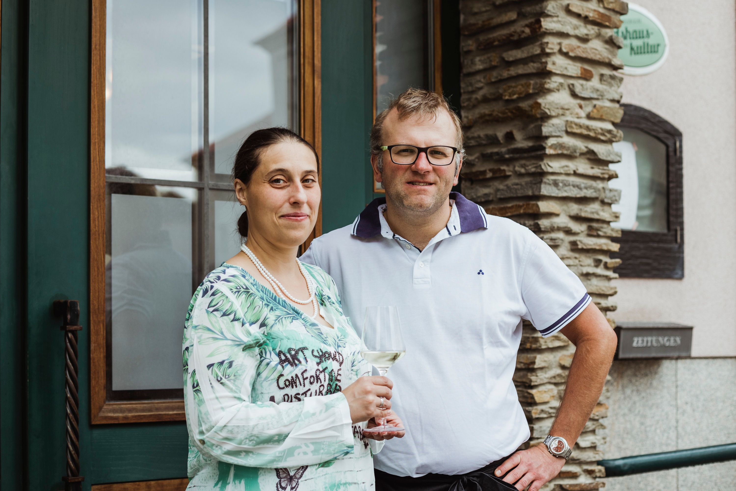 Egy férfi és egy nő áll mosolyogva egy épület előtt. A nő kezében egy borospohár.