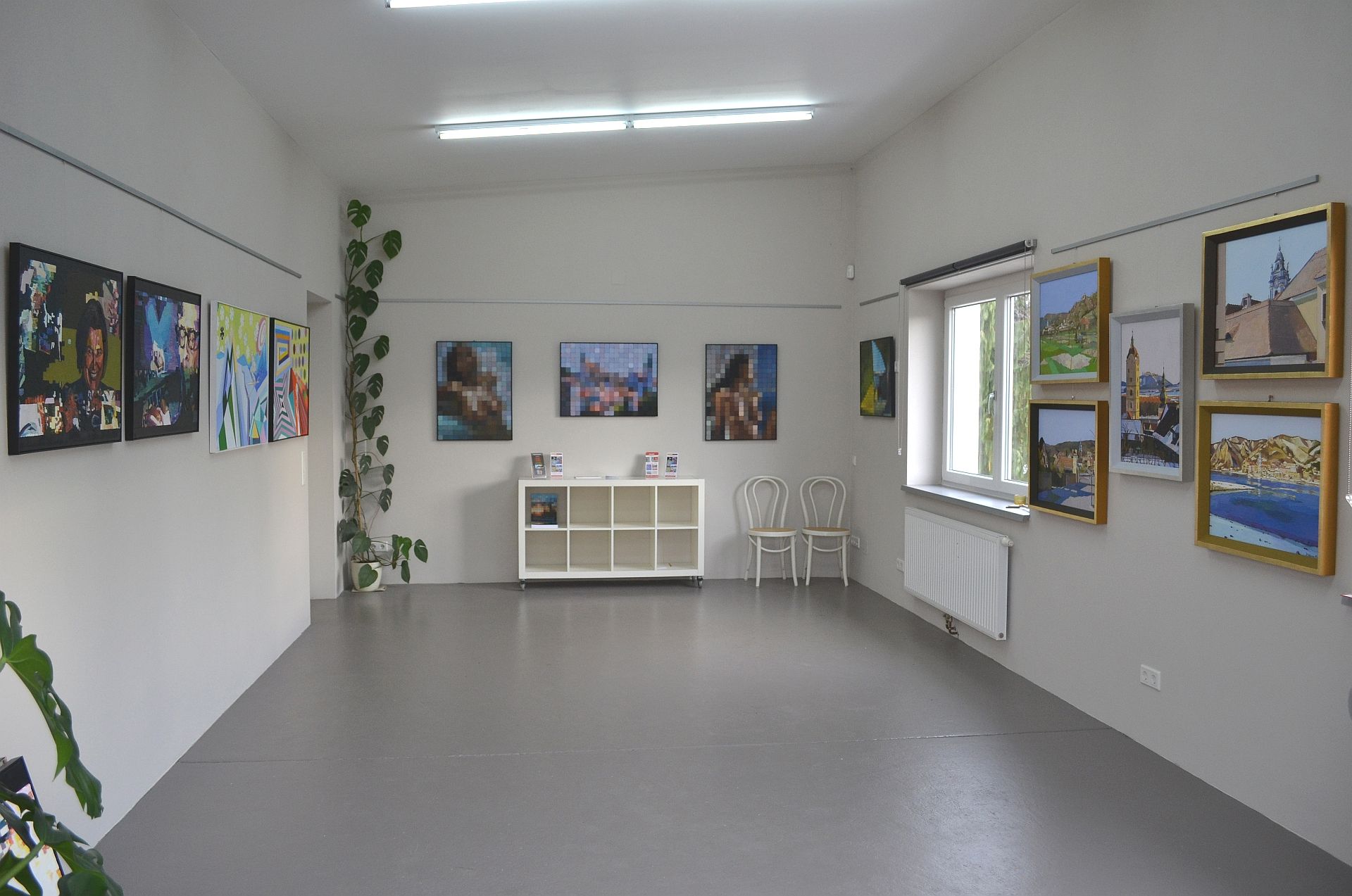 Egy galéria belső nézete, a falakon festményekkel, középen egy polccal.