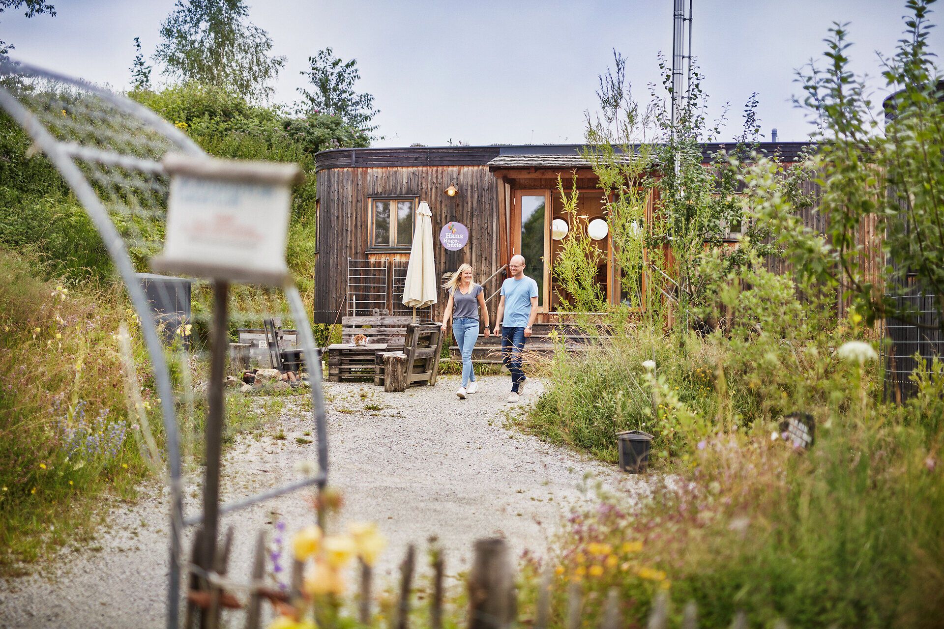 Ein Paar schlendert entspannt durch die blühenden Wiesen, umgeben von der idyllischen Landschaft. Die rustikale Holzarchitektur des Landlofts strahlt Gemütlichkeit aus und lädt dazu ein, die frischen Kräuter und die Natur in vollen Zügen zu genießen.