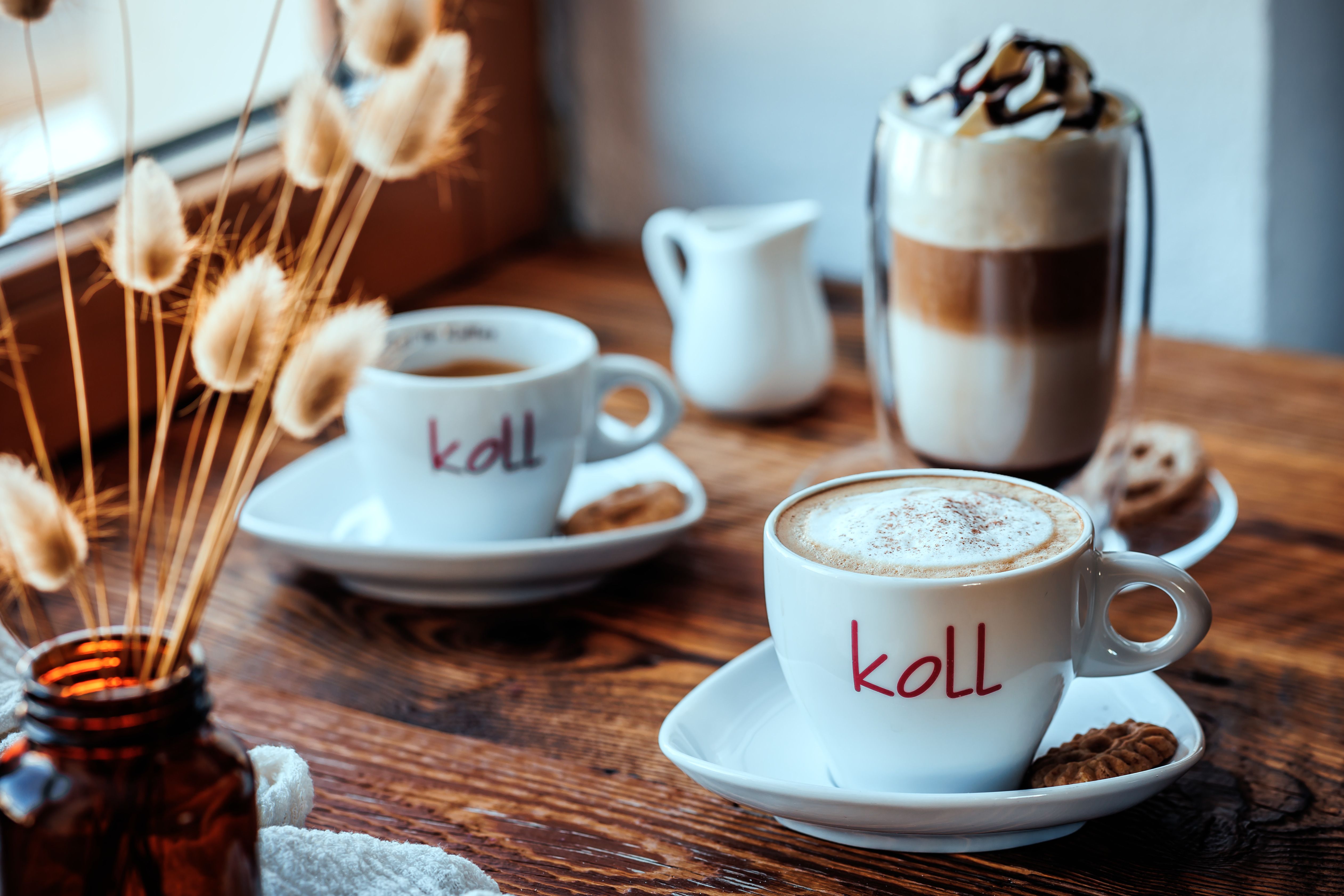 Két csésze kávé egy faasztalon, az egyikben tejhab, egy pohár latte macchiato és mellette egy kis tejeskanna.